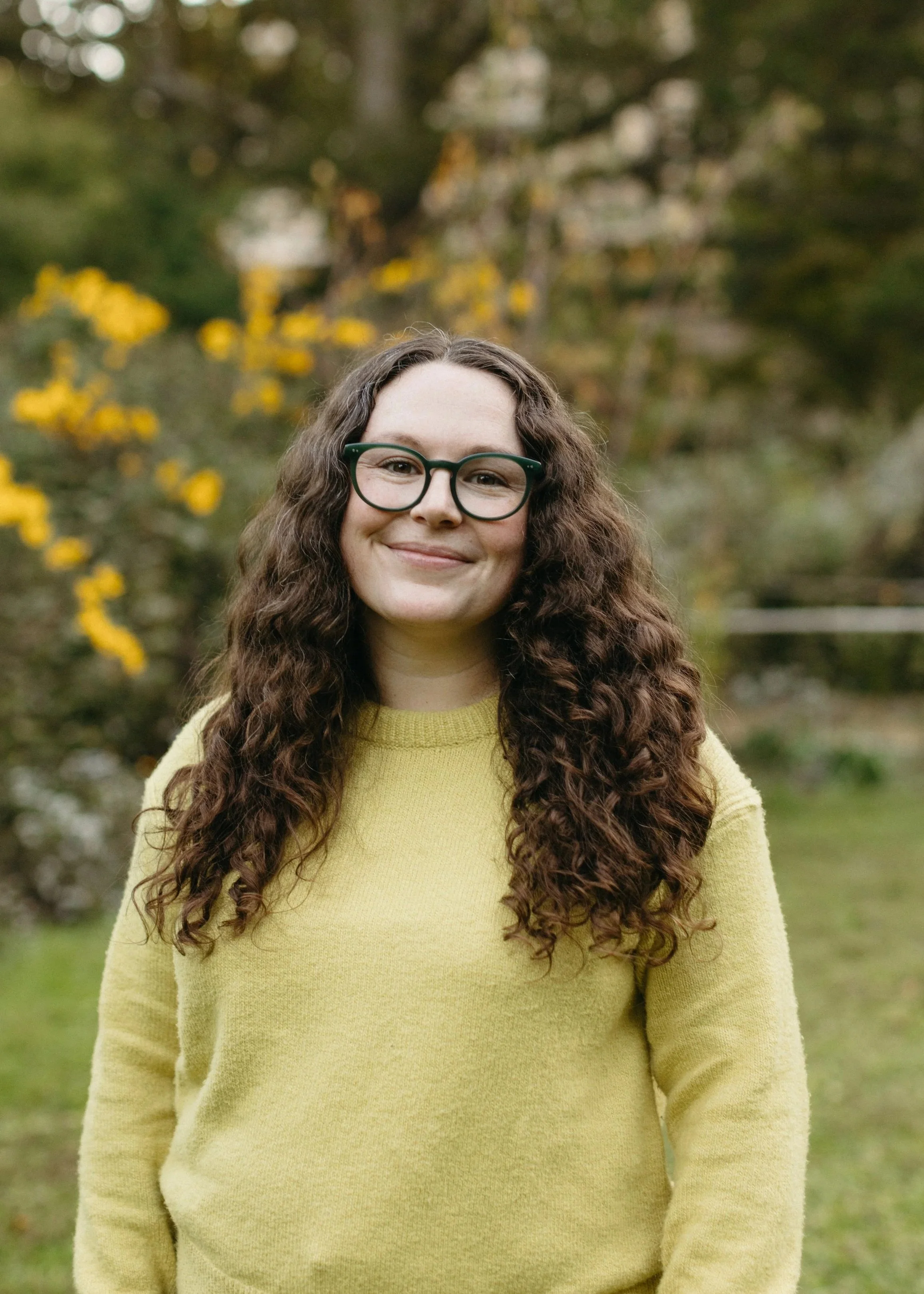 A woman with curly brown hair, glasses, and a yellow-green sweater standing outdoors with trees and yellow flowers in the background.