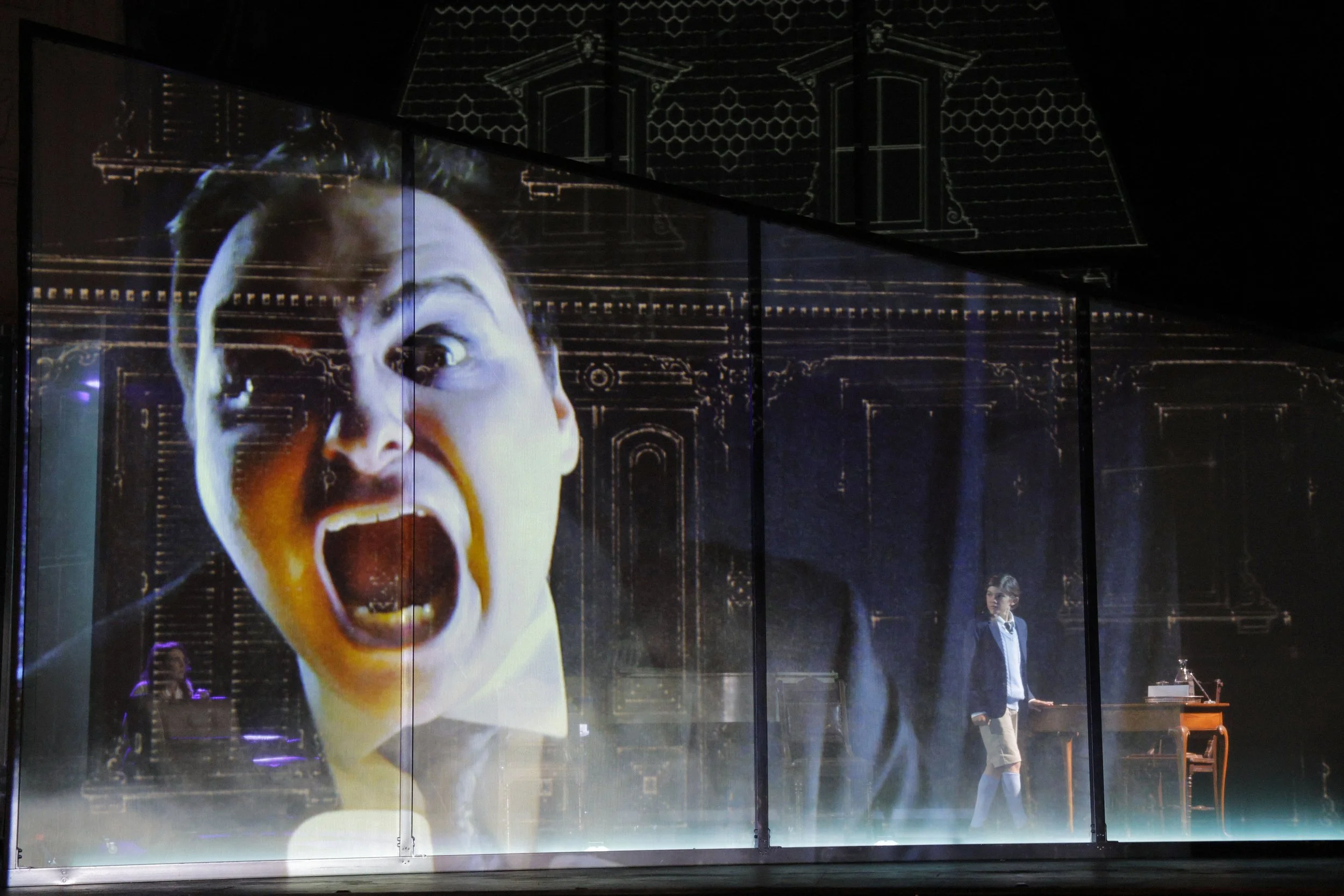 A large projection of a woman with a screaming expression, mouth wide open, on a dark stage or wall. In the background, a man in a suit is standing near a piano, and a smaller figure is visible near the bottom right.