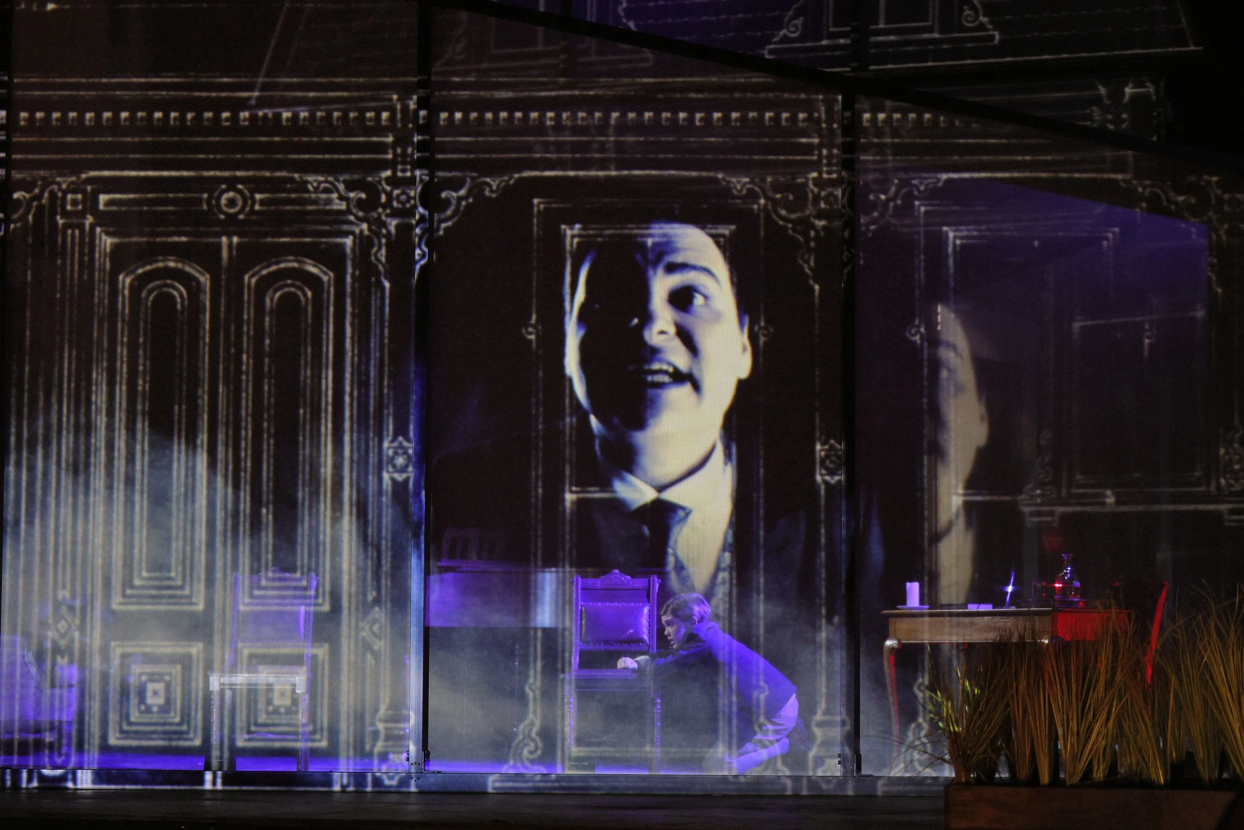 A theatrical stage with a large screen showing a black-and-white image of a man with a surprised expression. The stage has ornate wood paneling and is illuminated with purple lighting. A person is seen sitting at a table in the foreground.