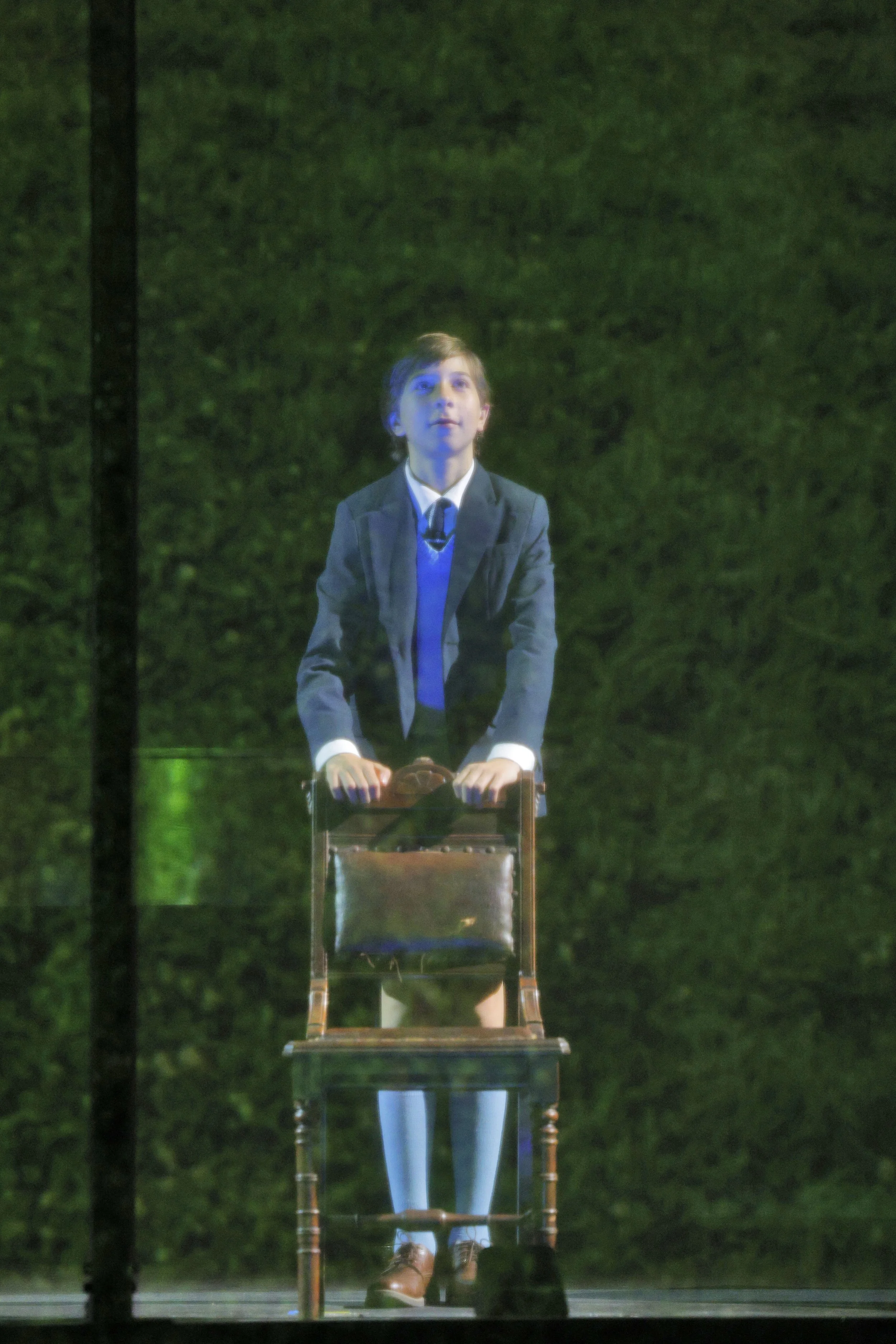 A young boy with brown hair dressed in a school uniform, standing behind a transparent podium with a green background.