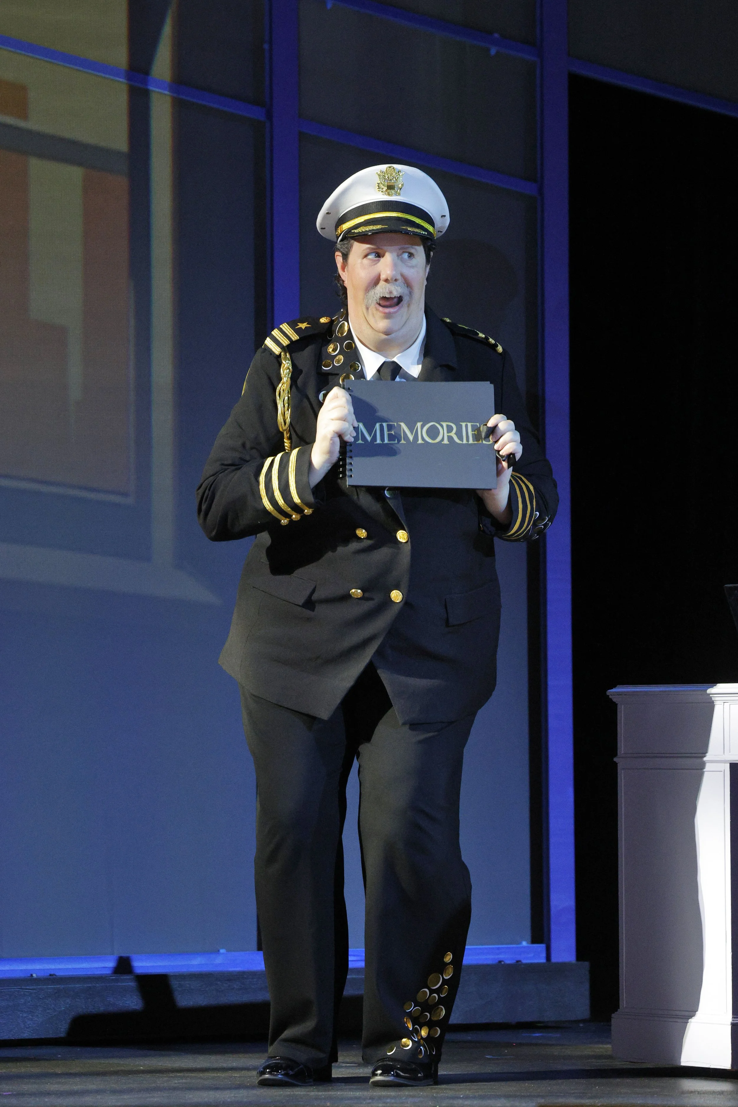 A man in a pilot's uniform holding a black folder labeled "MEMORIES," standing on a stage with a blue background and purple window frames.