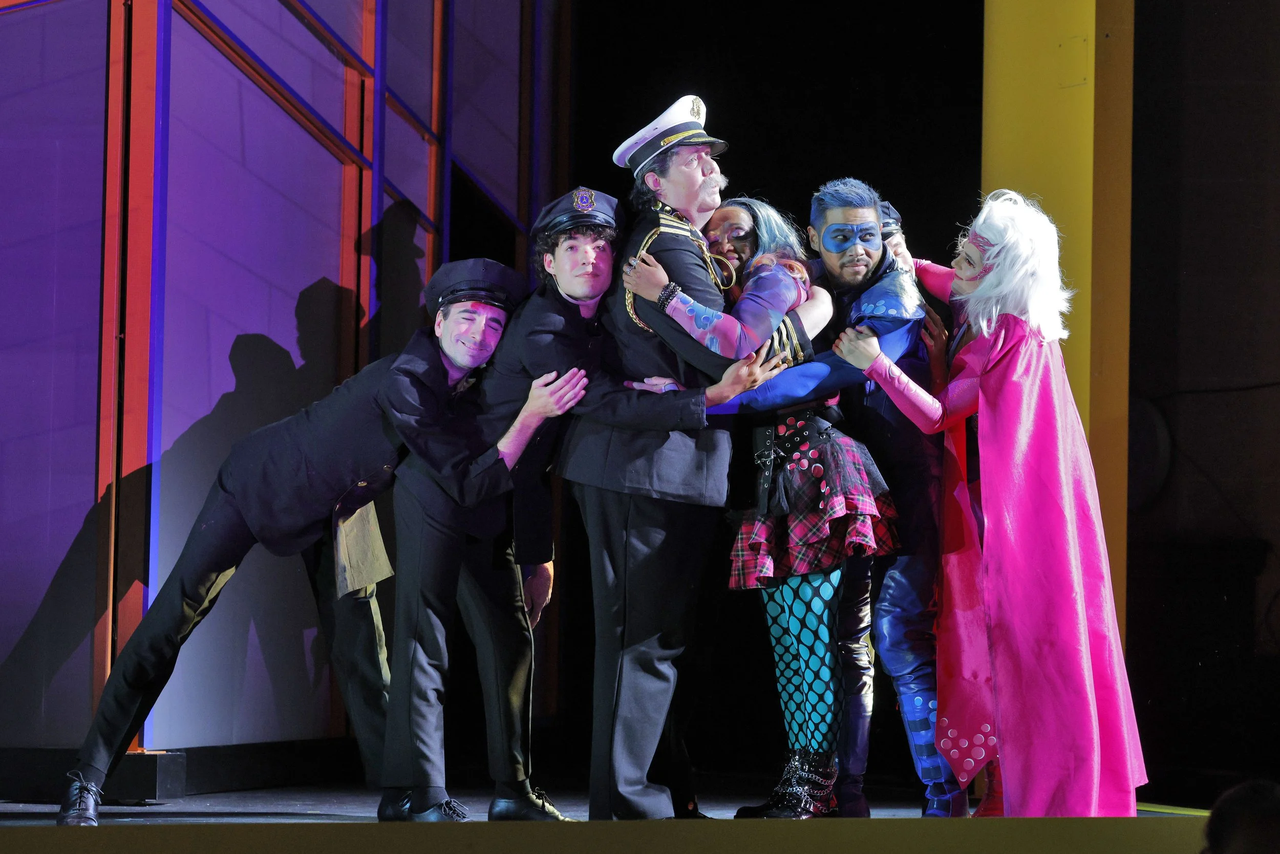 A group of performers in colorful costumes and makeup, hugging and smiling on stage during a theatrical performance.
