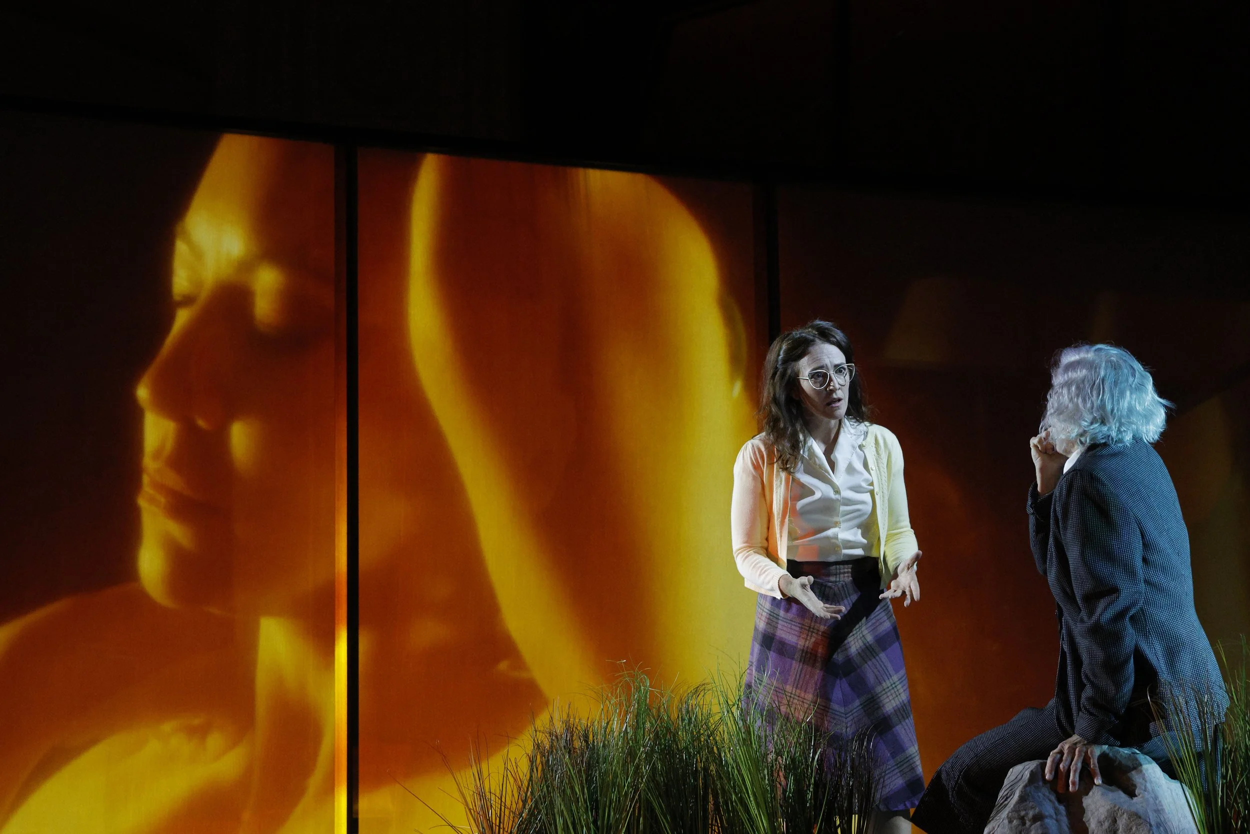 Two women speaking on stage with a vibrant, orange-yellow abstract background projection.