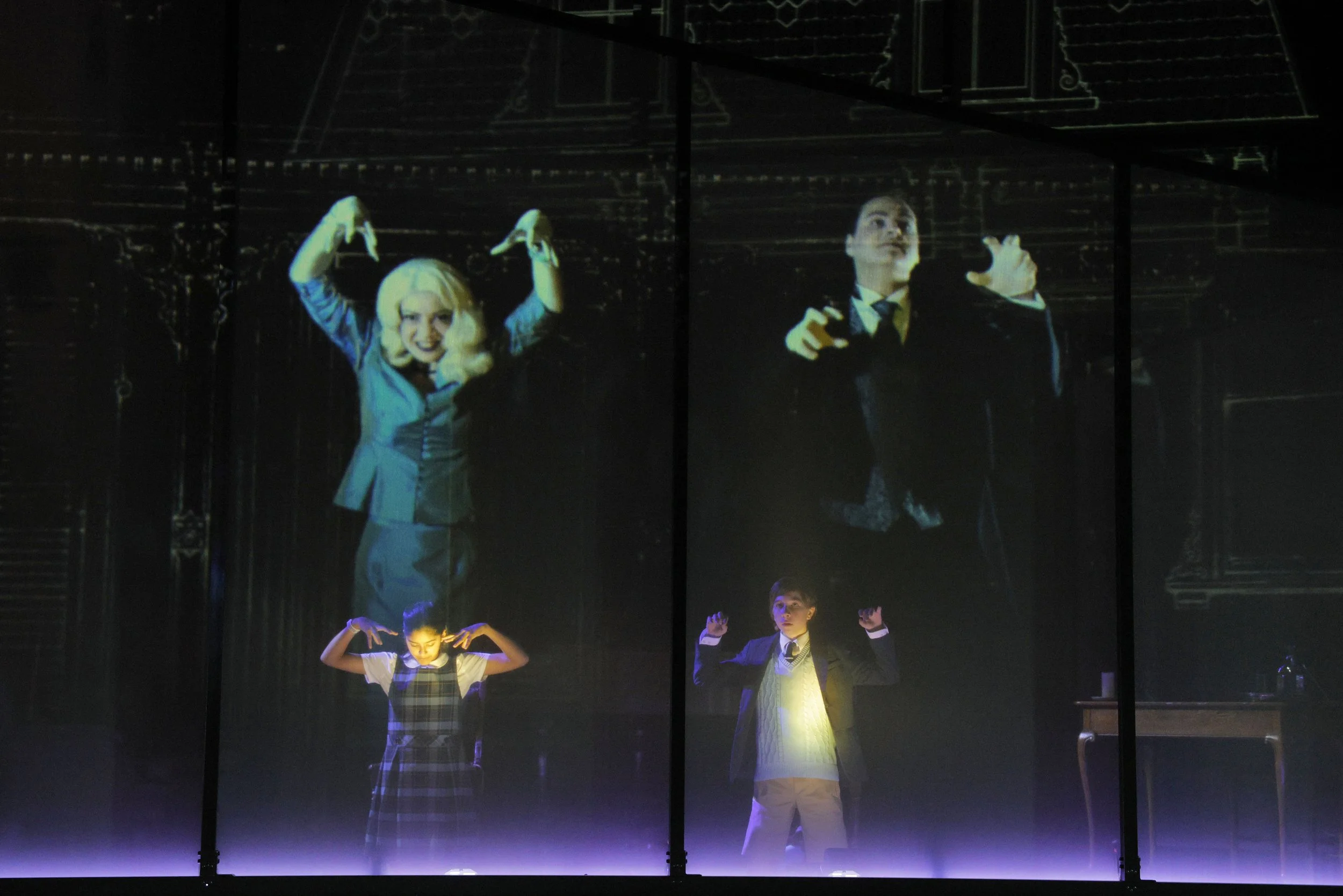 Stage scene with projected images of actors and children performing, some with hands raised and making gestures, in a dark theater with minimal lighting.