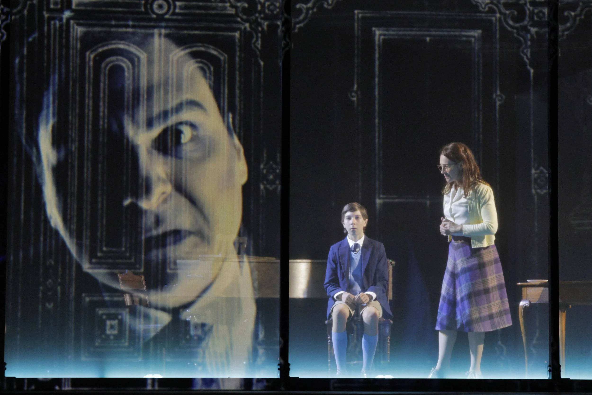 The image shows a scene from a theatrical play featuring three characters, two children dressed in school uniforms, and an adult woman, all standing on stage in front of a large backdrop with a painted portrait of a woman with a solemn expression. Th
