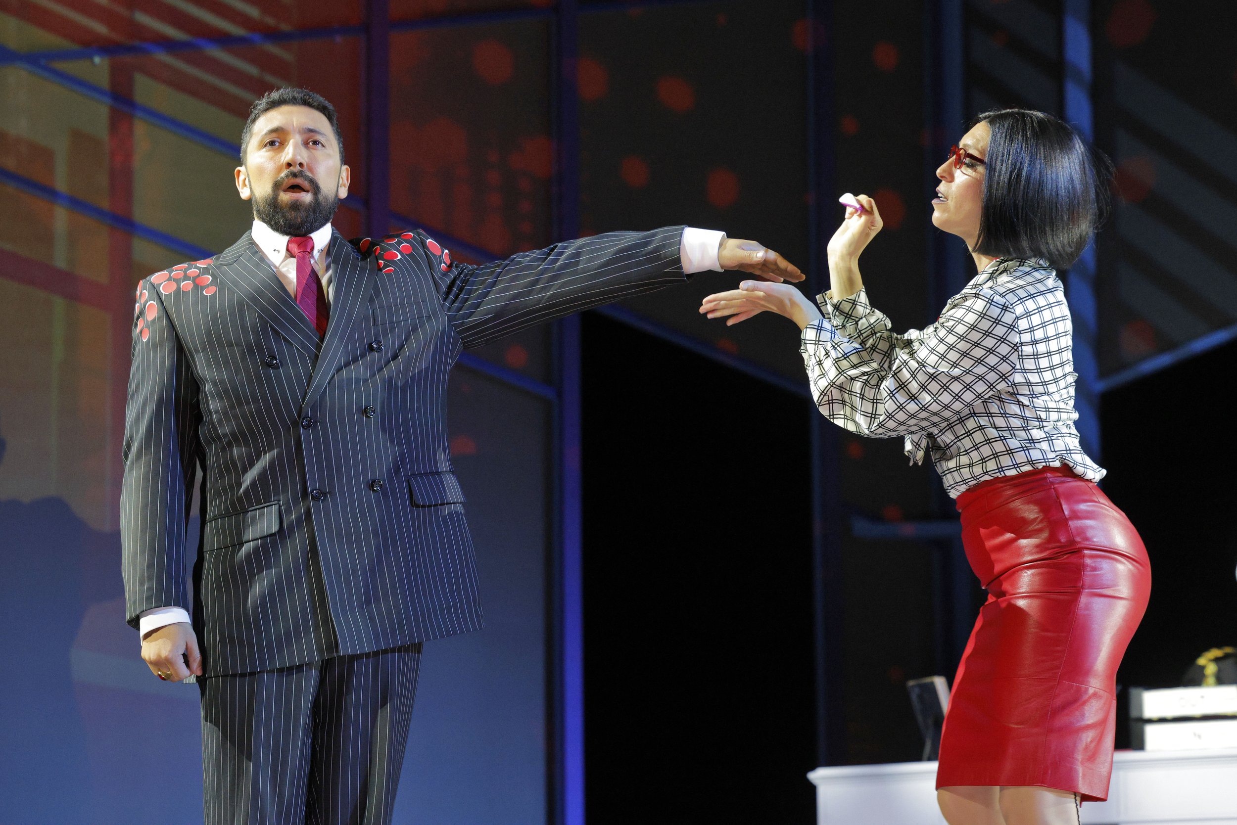 A man with a beard dressed in a pinstripe suit with a red tie is reaching out to a woman with black hair, glasses, wearing a white and black checkered top and red leather skirt, on a stage with a dark background.