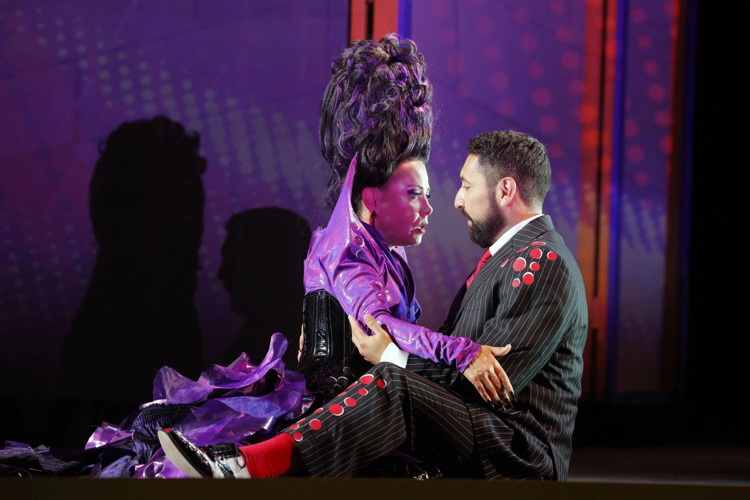 Two actors, dressed in colorful costumes, are on stage close together, gazing into each other's eyes. The female actor has elaborate hair and makeup, wearing a shiny purple outfit with purple ruffles. The male actor has a beard, short hair, and is dr