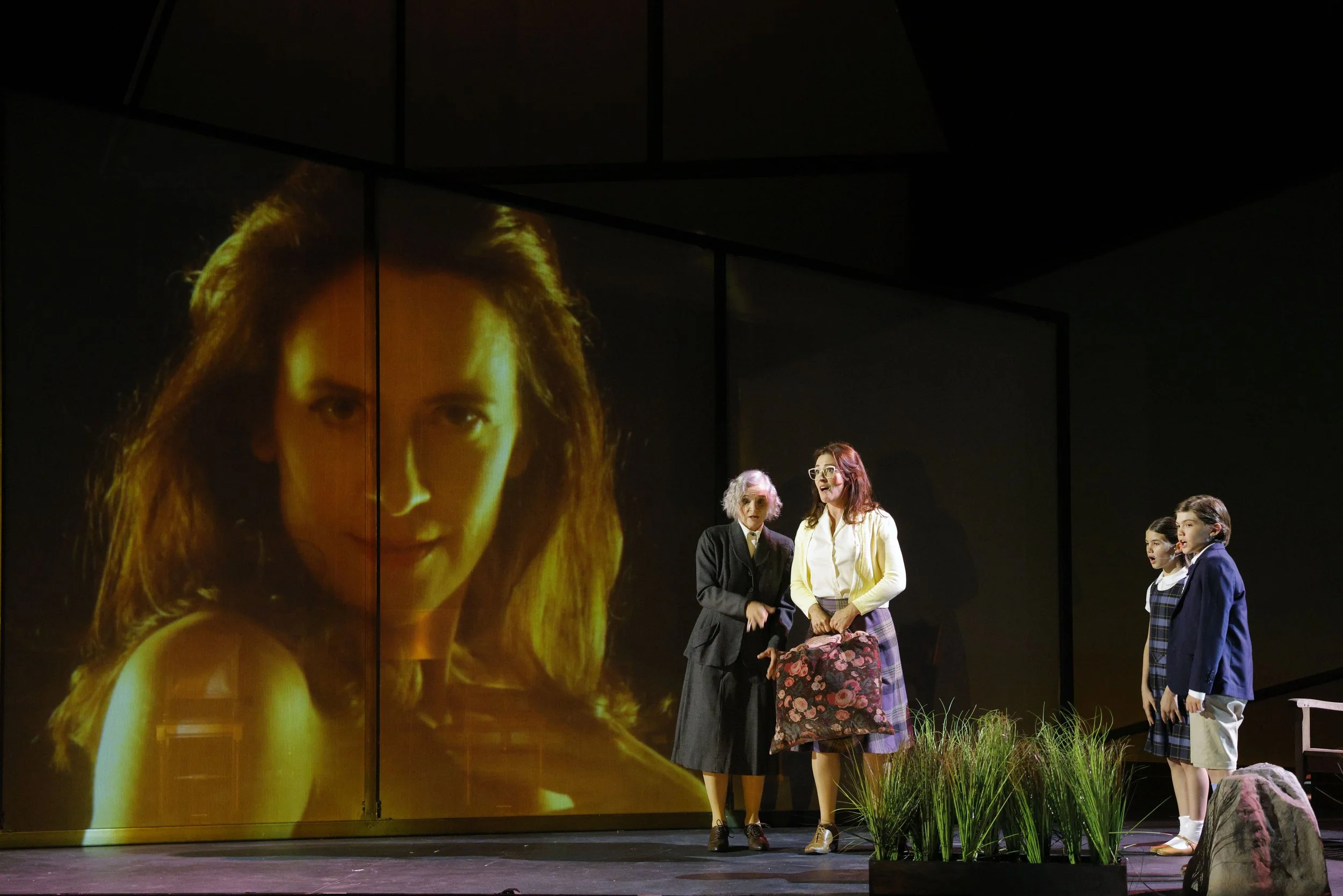 Stage scene with five women and four girls in school uniforms, with a large projected image of a woman with long hair in the background.