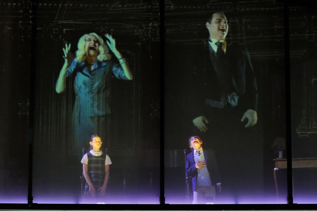A scene from a stage play with two large projected images and two children in school uniforms. The woman on the left appears to be shouting or screaming with hands raised, and the man on the right has a stern expression with hands on his hips. The ch