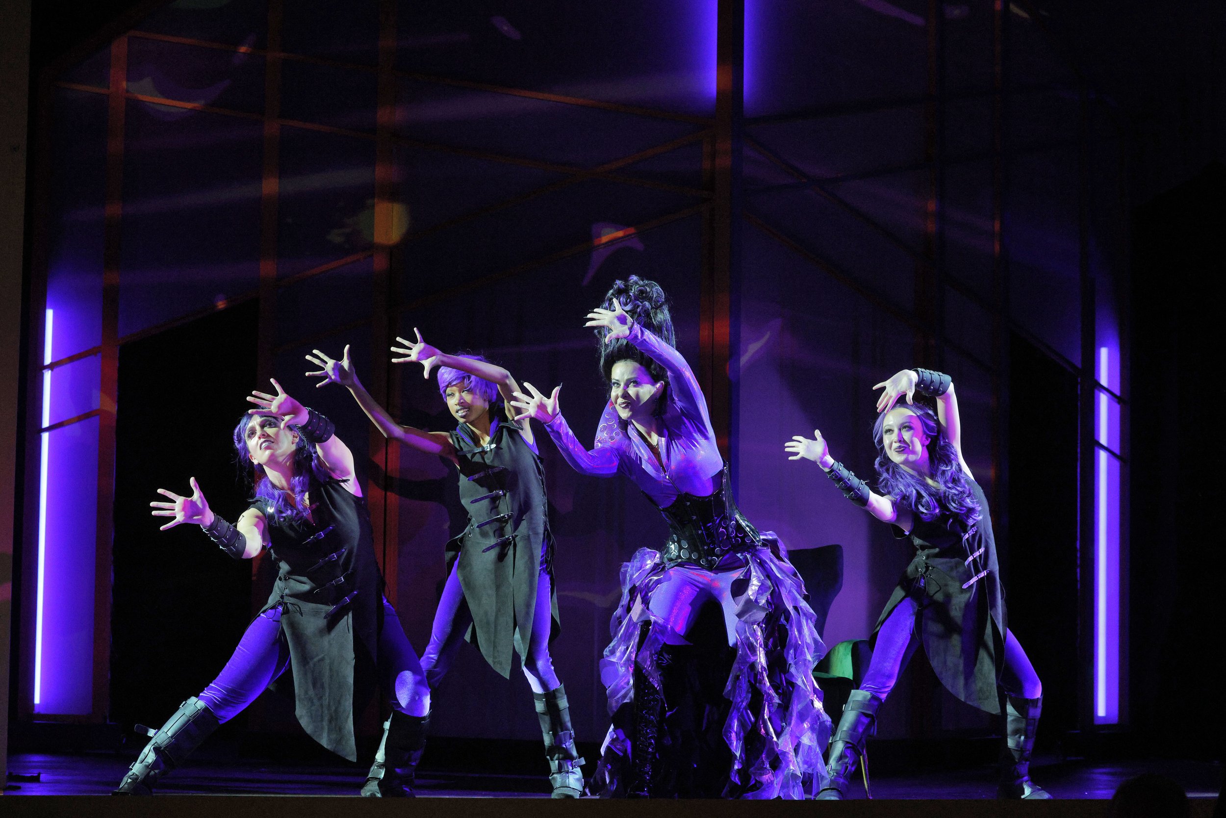 A group of five performers in colorful costumes on stage with purple and blue lighting, striking expressive poses during a theatrical dance or performance.