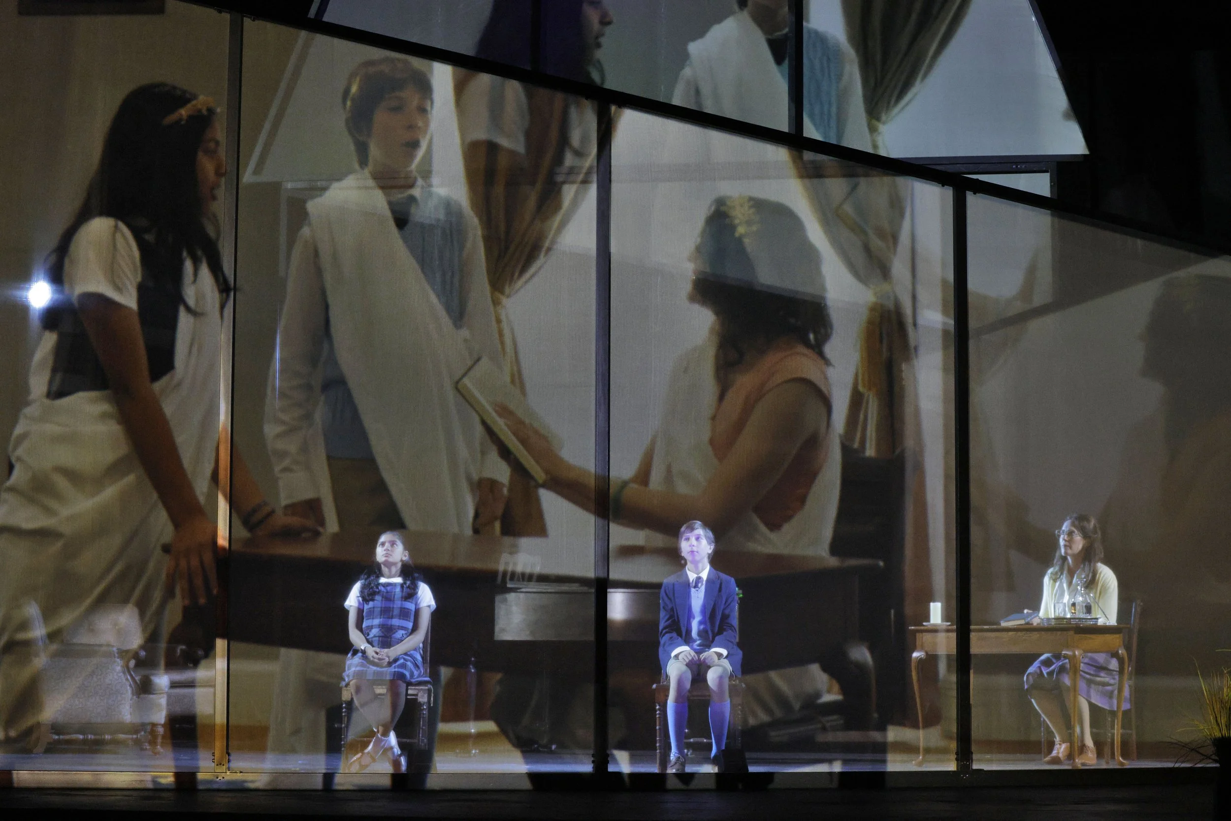 People gathered around a table with a glass display case showing miniature figures, with reflections of individuals watching from behind.