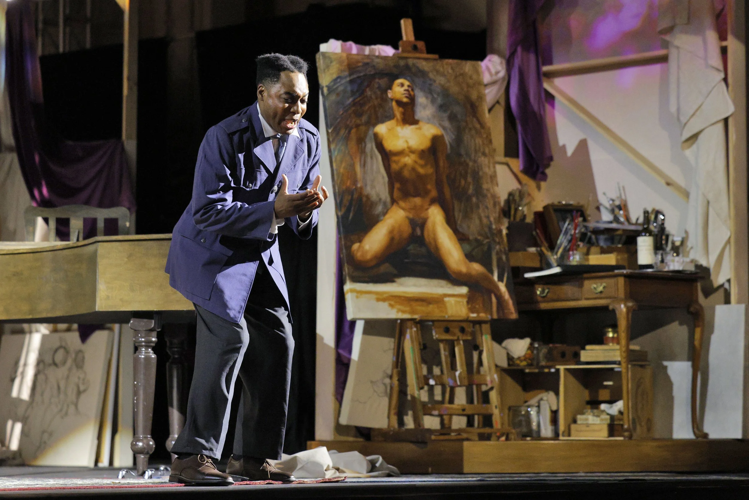 A man in a blue suit standing on a stage, gesturing with his hands, with a painting of a nude woman on an easel behind him. The background shows art supplies and purple curtains.