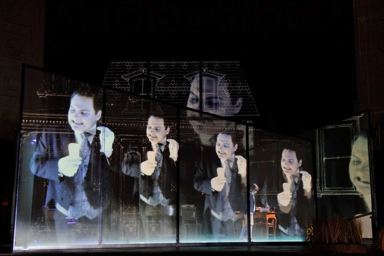 Multiple screens displaying a man in a black suit holding a microphone, with his image reflected on glass panels.