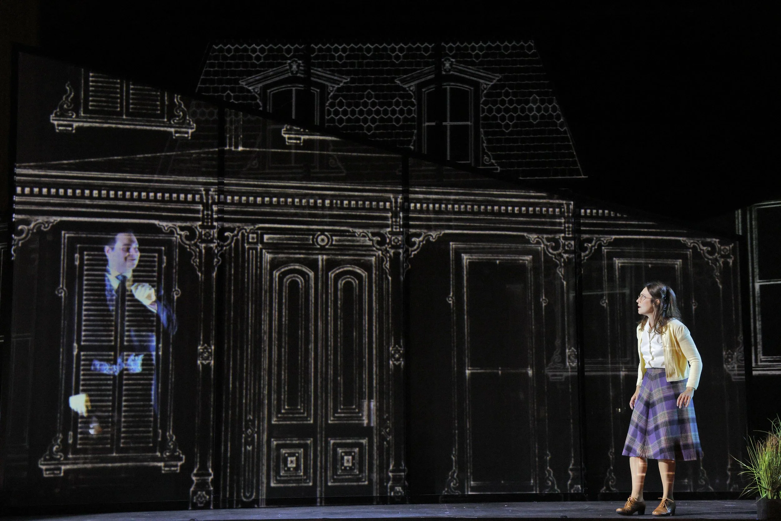 A woman with long dark hair and glasses walking past a building with a large projection of a man in a suit and a woman in a plaid skirt.