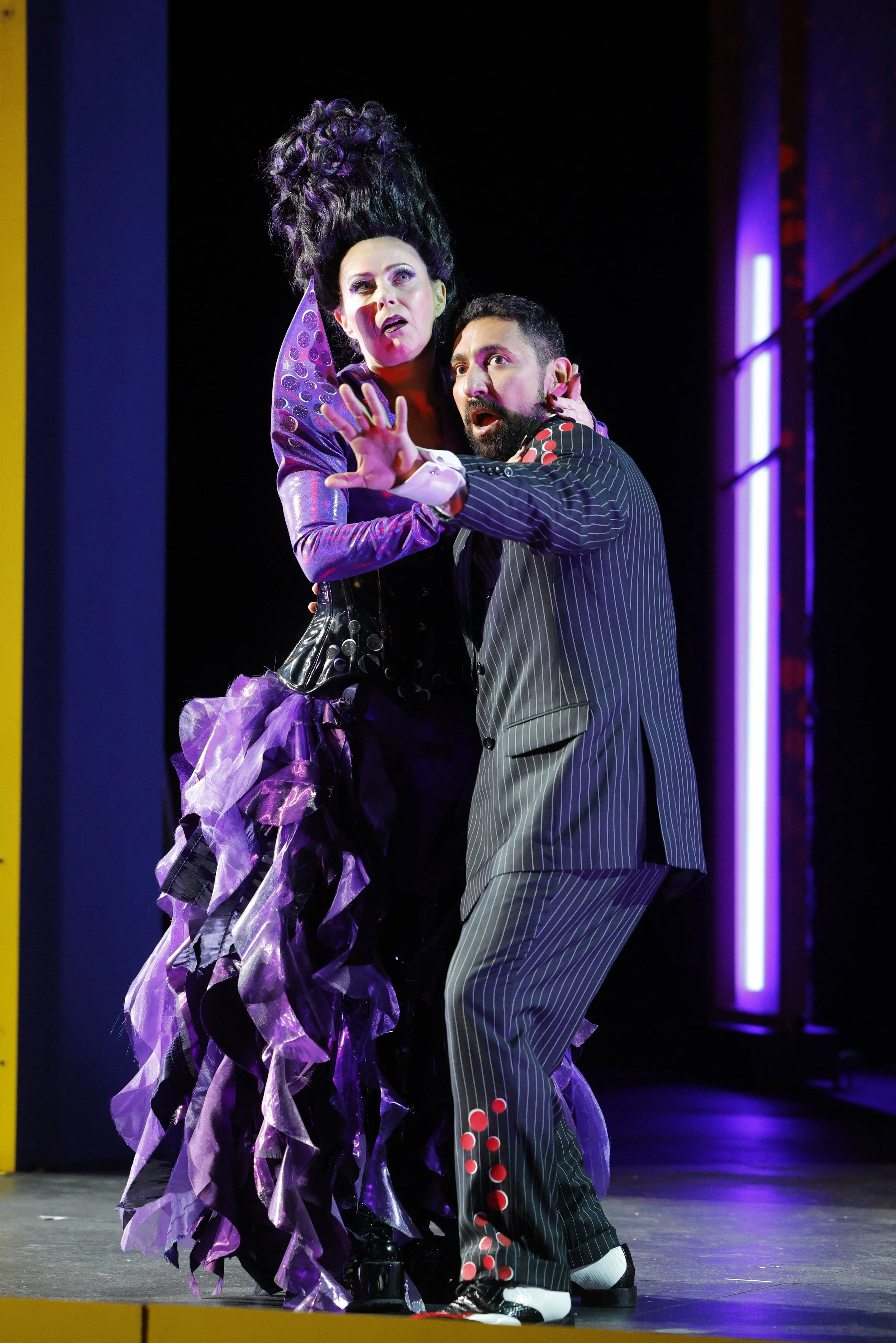 Two performers on stage, one with tall dark hair and elaborate purple costume, the other with short dark hair and striped suit, appearing surprised or alarmed.