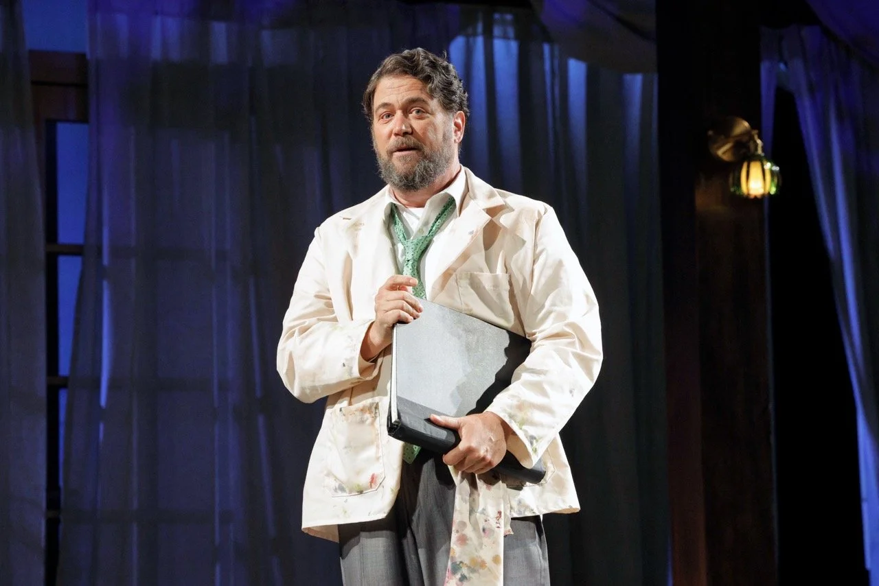 A man with a beard and curly hair, dressed in a white coat, holding a laptop, standing on a stage with dark curtains and warm lighting.
