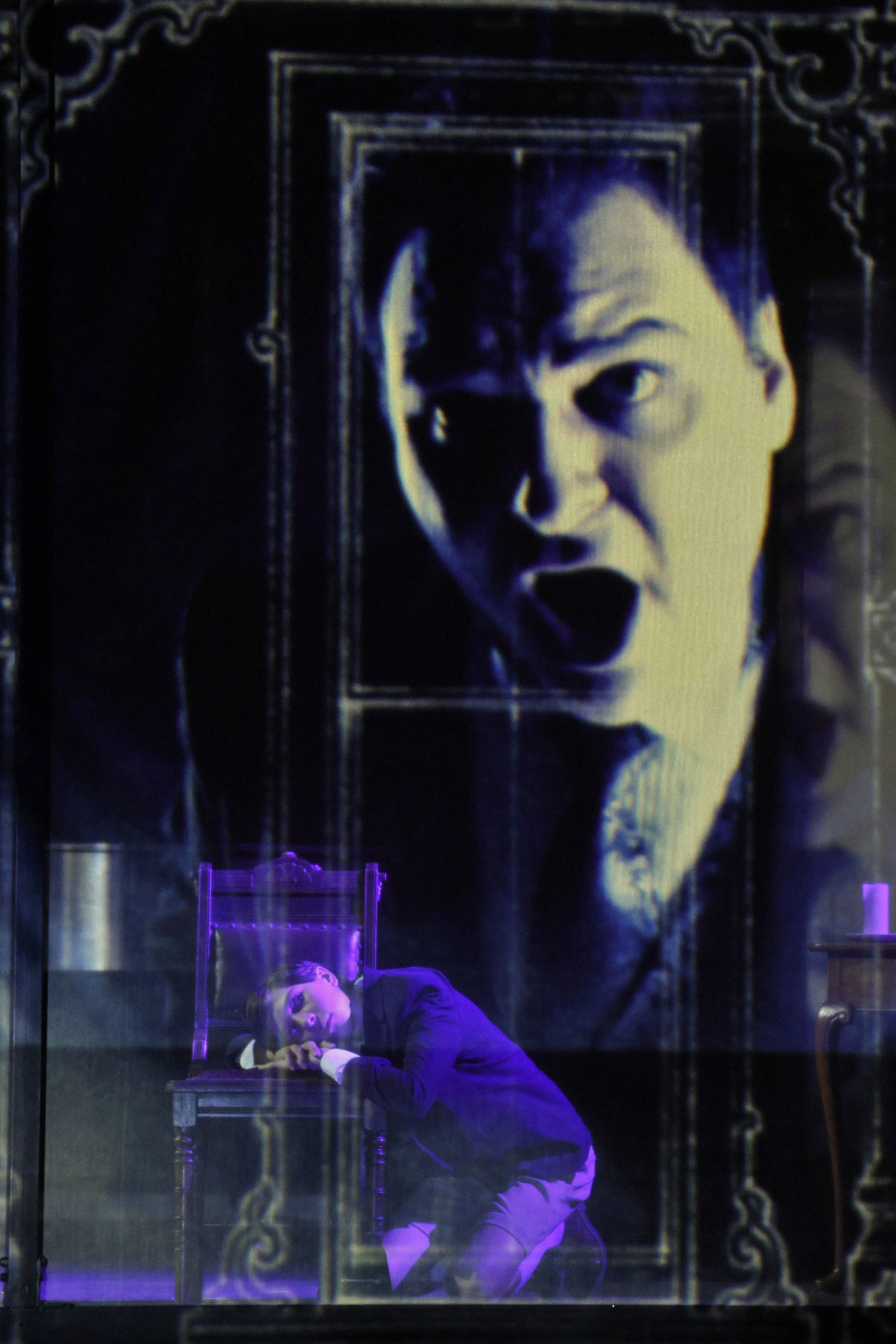 The image features a person lying on a table, gazing upwards, with their hands clasped together under their chin. Behind them is a large, black-and-white portrait of a young woman with an expressive, open-mouthed facial expression. The scene is lit w