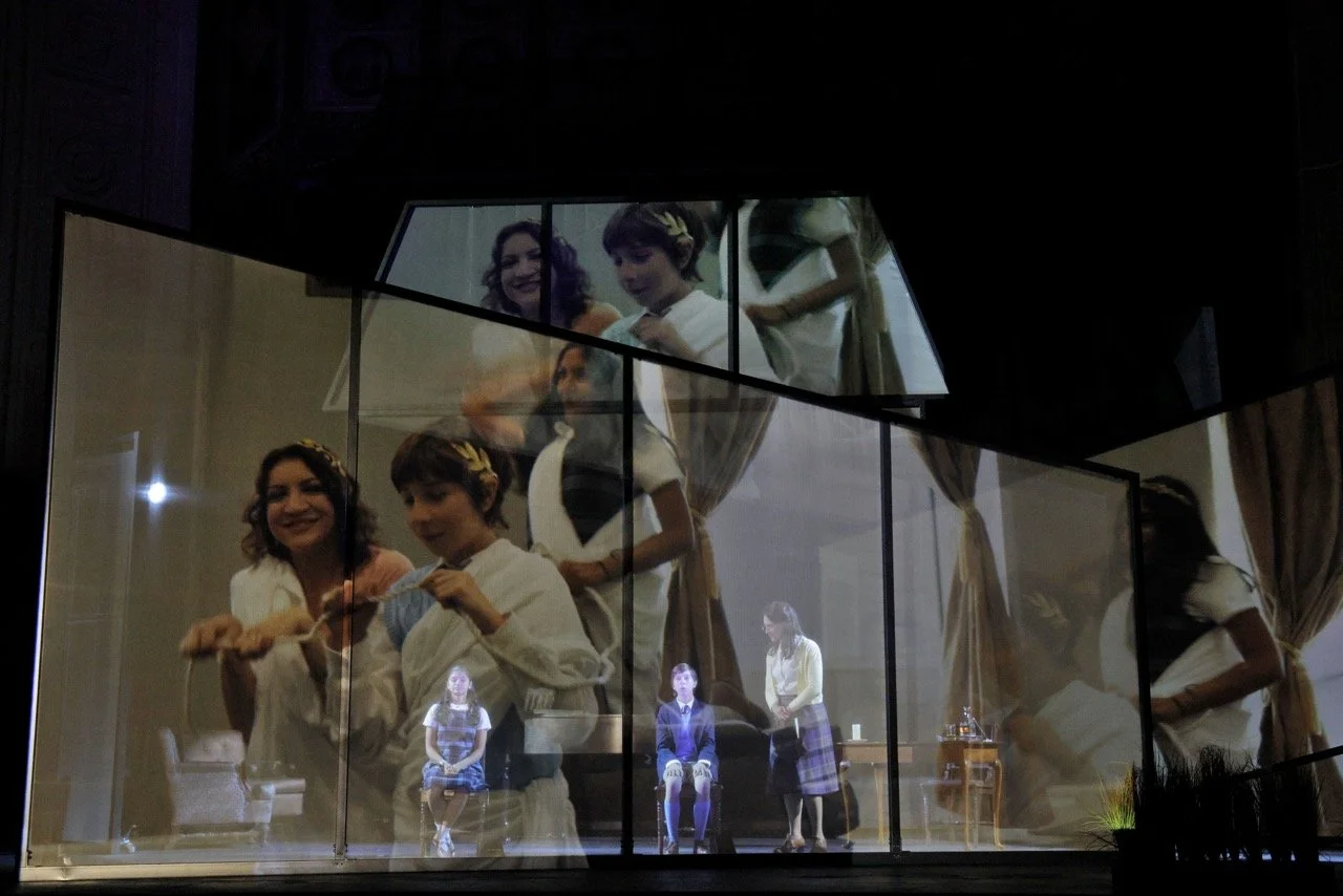 The image shows a theatre scene with actors performing under a backdrop that reflects their images, creating a layered visual effect.