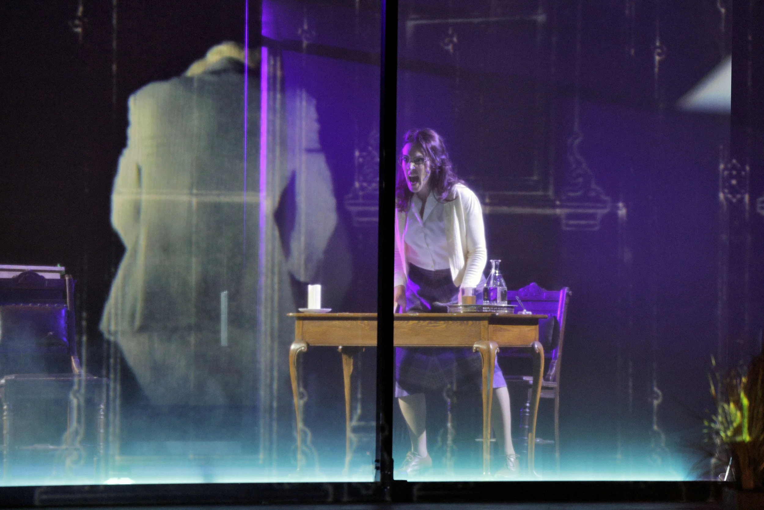 Scene viewed through glass, featuring a woman with glasses and curly hair, standing behind a table with a water pitcher and glass. The woman appears to be shouting or reacting emotionally, with a dark background and purple lighting.