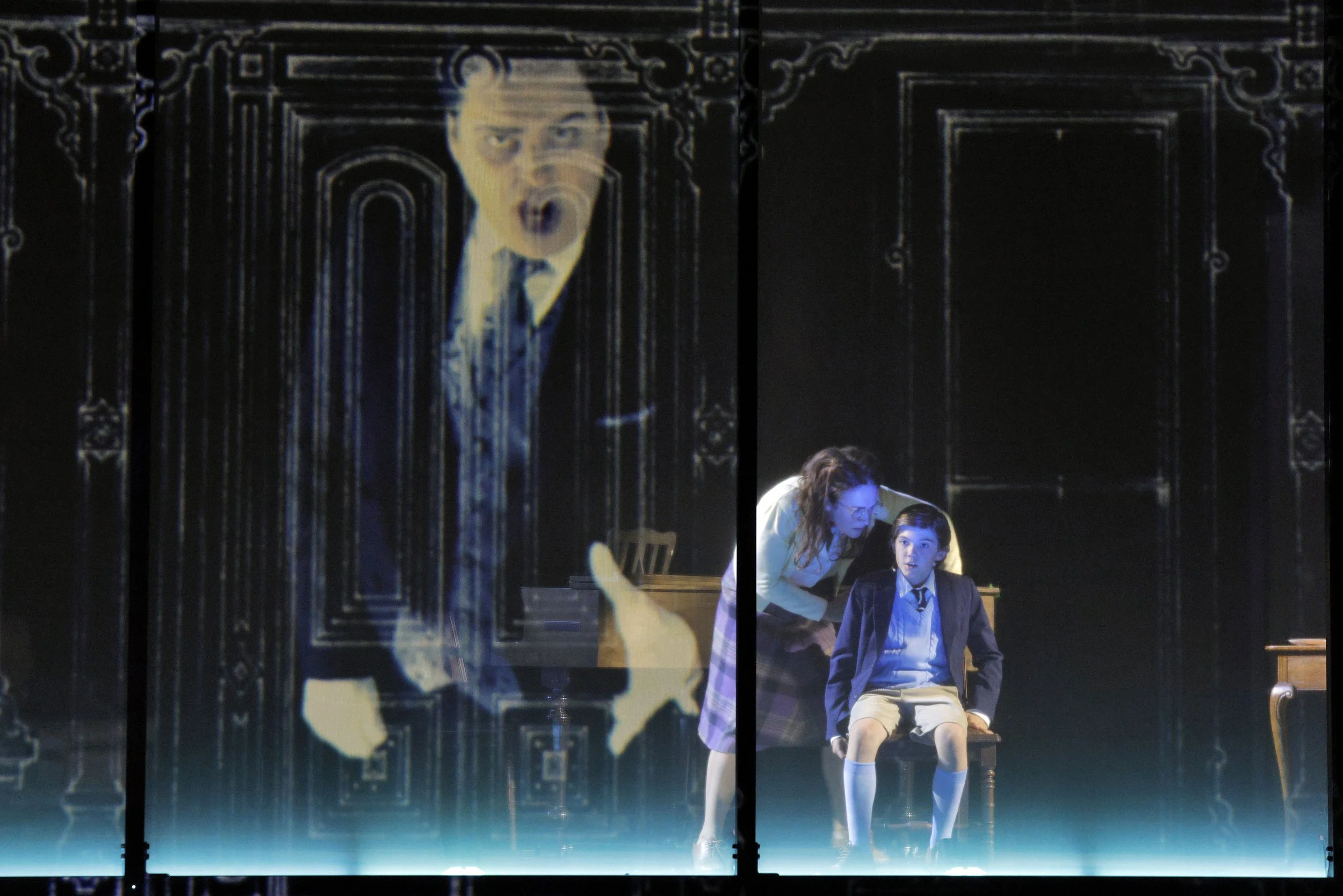 The image shows theater scenery with painted dark wood doors and ornate framing. In front, a woman in a beige jacket and glasses is seated with a young boy in a school uniform, sitting on a bench, engaged in conversation. A projected image of a surpr