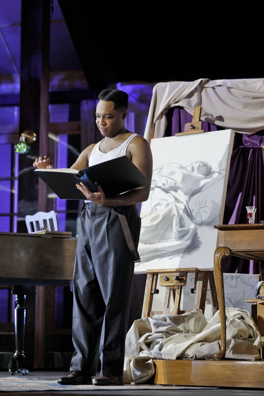 A person standing and reading from a large book on stage, with a drawing of a reclining nude figure on an easel behind them.