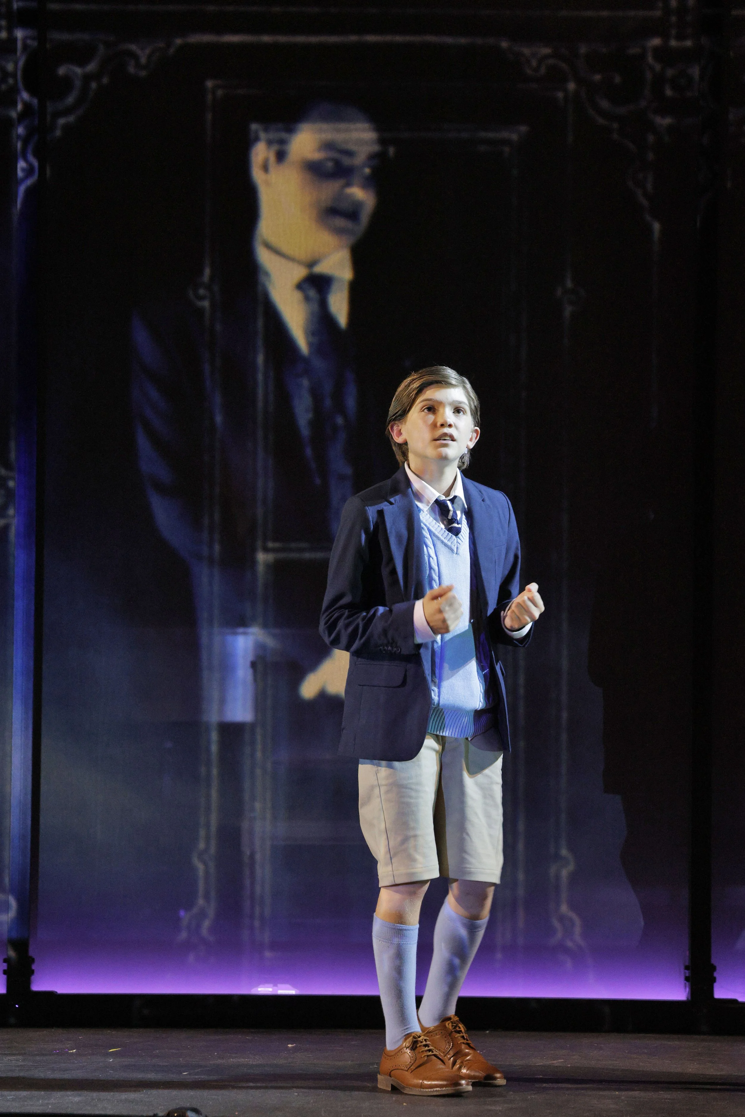 A young male student in a school uniform, including a blazer, tie, sweater, and khaki shorts, standing on stage with a microphone in his hand, in front of a large screen showing a woman's face.