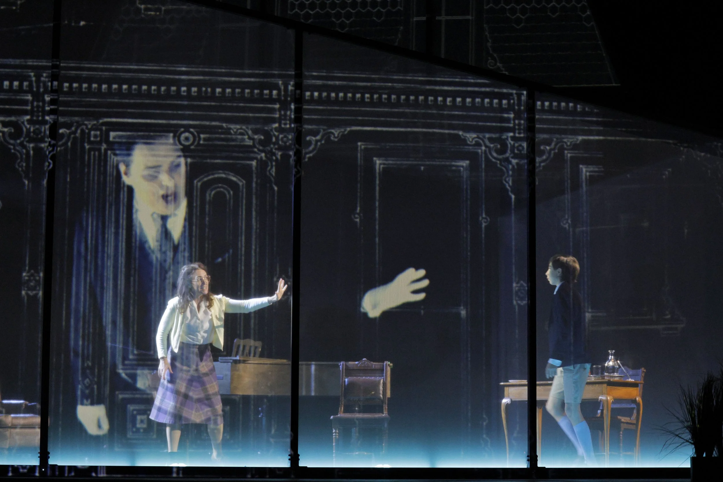 The image shows a stage scene from a play or performance, viewed through a glass with black framing. There are three actors on stage: a woman with long, curly hair in a white blazer and plaid skirt, gesturing with her arms and speaking; a woman with 