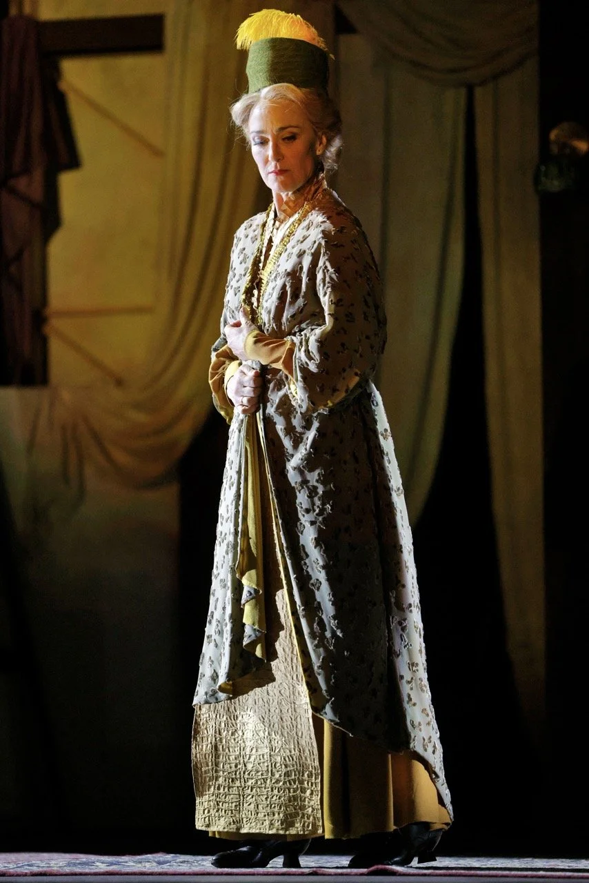 A woman dressed in an elaborate costume, standing on stage with a serious expression, wearing a tall green and yellow hat, vintage-style clothing with gold accents, and a patterned robe.