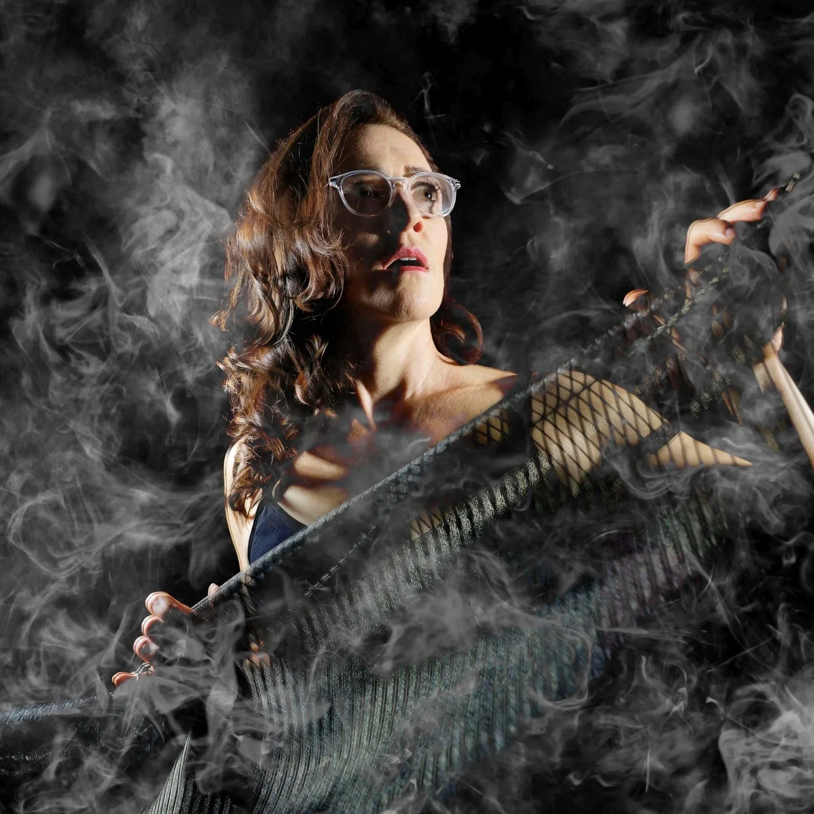 A woman with wavy brown hair, wearing glasses, a black fishnet top, and striped pants, is surrounded by smoke or fog and holding a baseball bat.