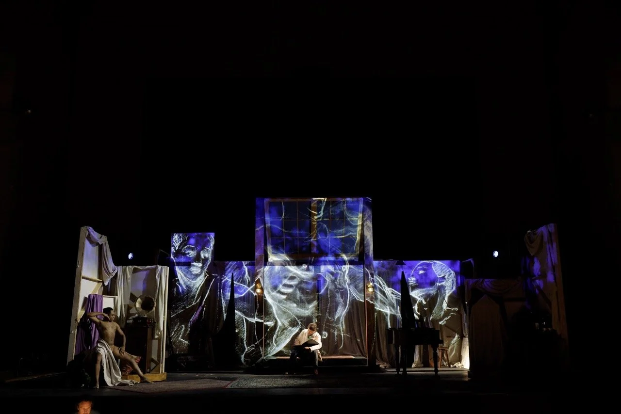 Stage set with dark background, curtains, and dramatic projection of faces on screens; actors sit or stand in period costumes, suggesting a theatrical performance.