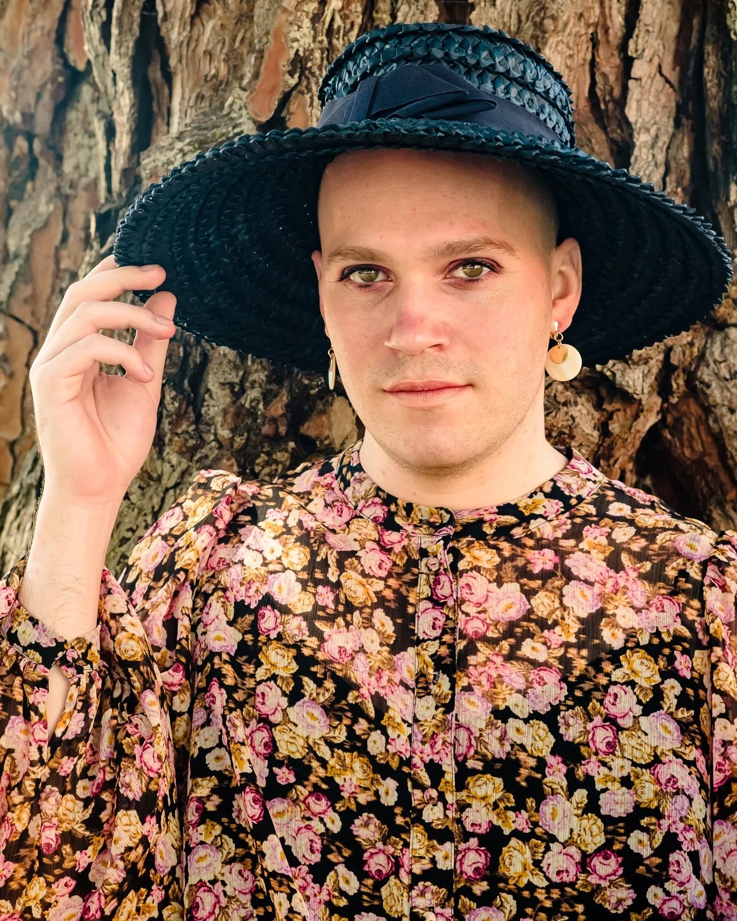 Person with short hair wearing a large black hat with a ribbon, a floral patterned shirt, and earrings, standing in front of a tree trunk.