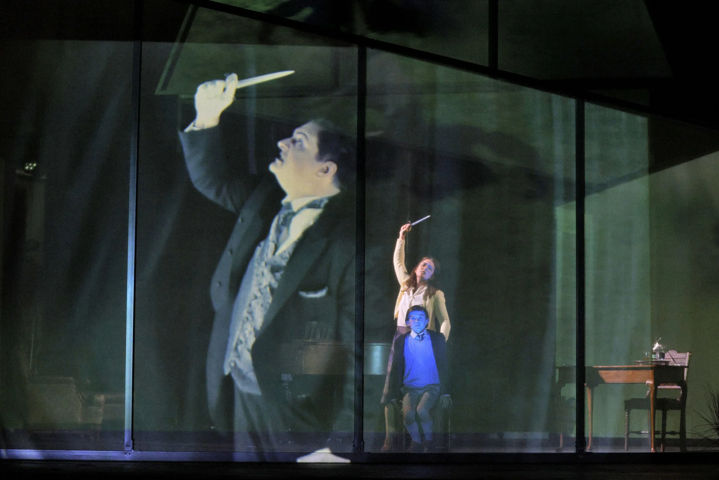A projected image of a conductor with glasses and striped tie conducting an orchestra, with three children in front of the projection, one girl raising her hand and two boys standing below.