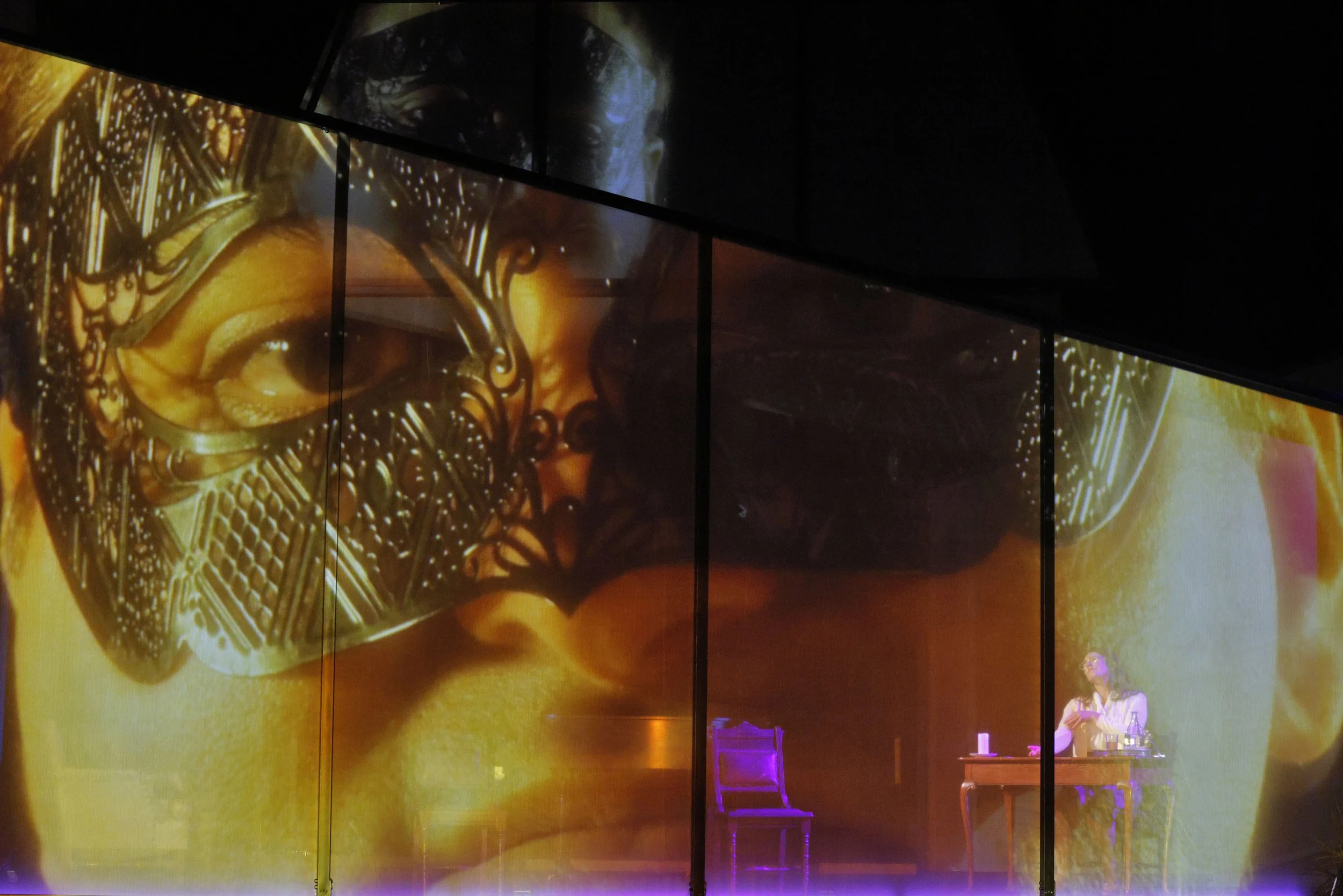 Large multimedia art display with vibrant, detailed images including a person's face, intricate black and gold patterns resembling a mask or decorative design, and a color gradient background with purples, oranges, and yellows. A woman stands on the 