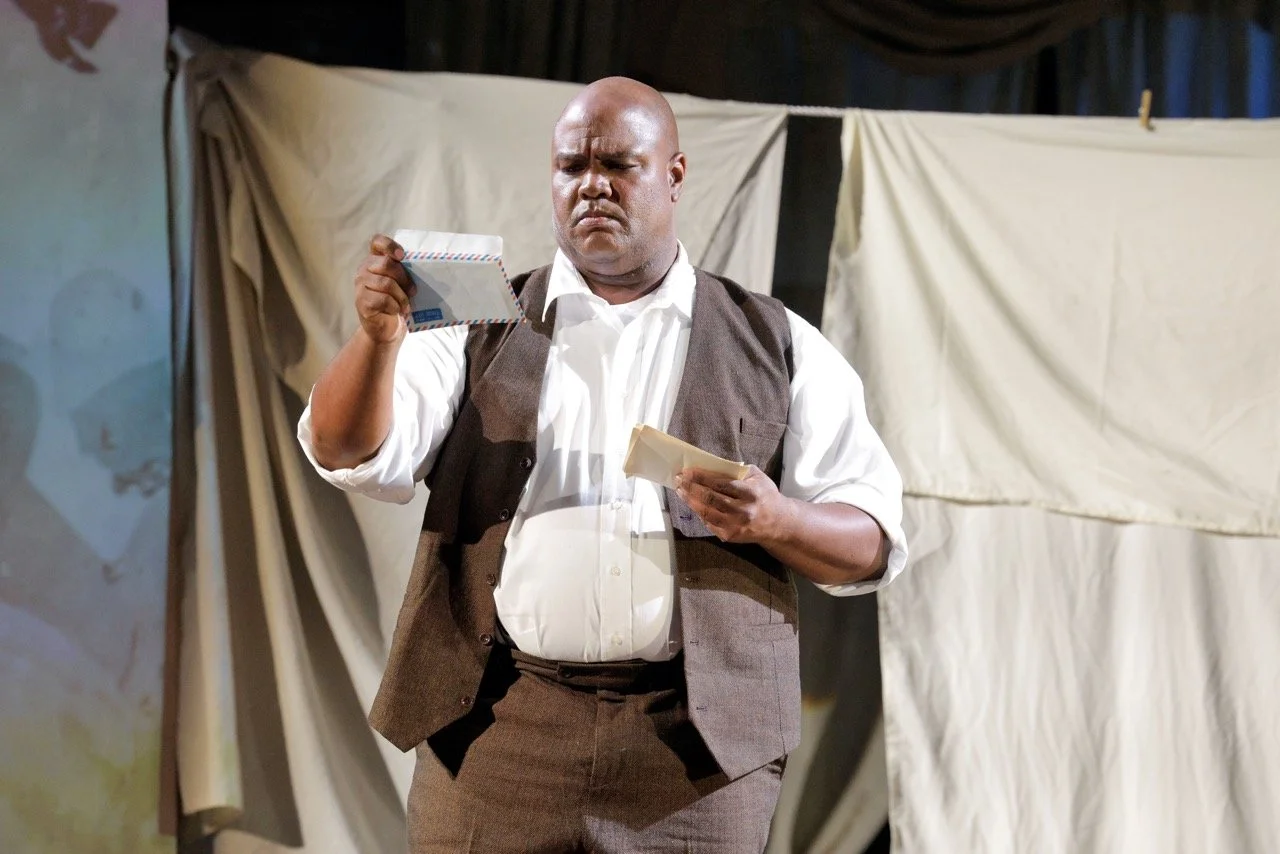 A man dressed in a white shirt and brown vest looking at mail in his hands on stage with a backdrop of fabric.