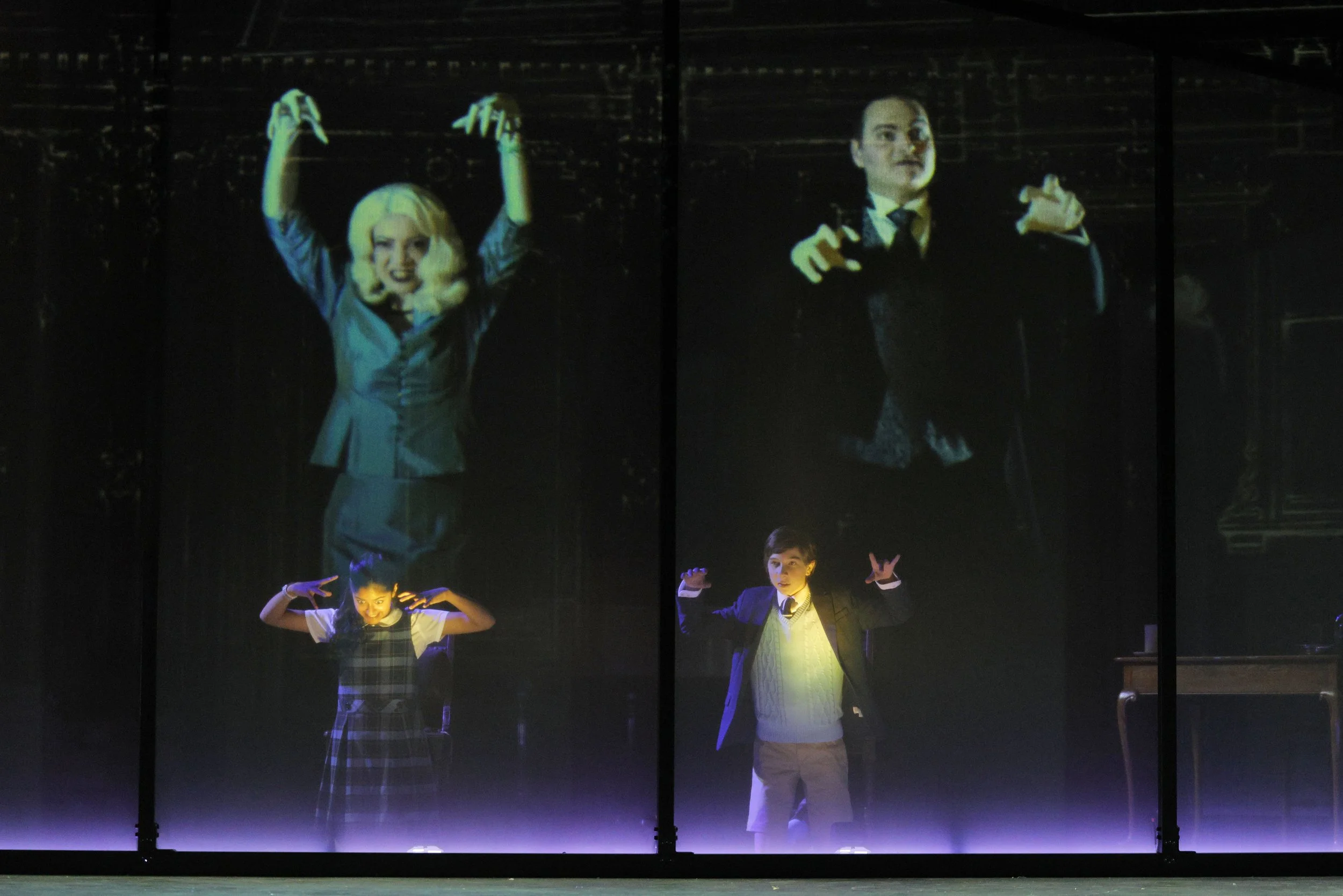Four children performing on stage under colorful lighting: a girl in a plaid dress in the front left, a girl in a school uniform on the front right, and two women in costumes behind them, one with blonde hair and the other with dark hair.