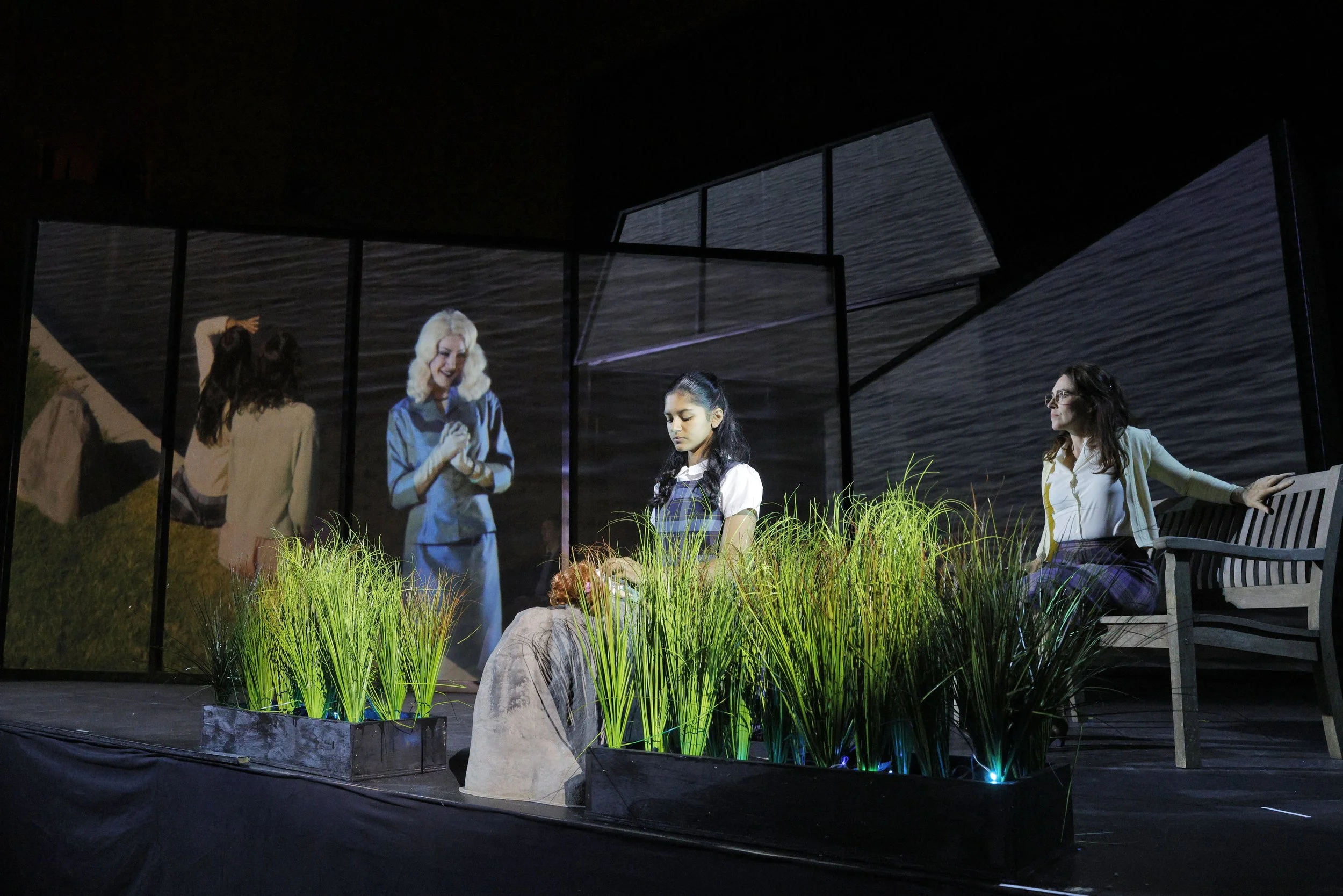 Nighttime indoor scene with four women, some with long dark hair and one with white hair, interacting near a window with water view. There are green plants in the foreground, and a black bench to the right.