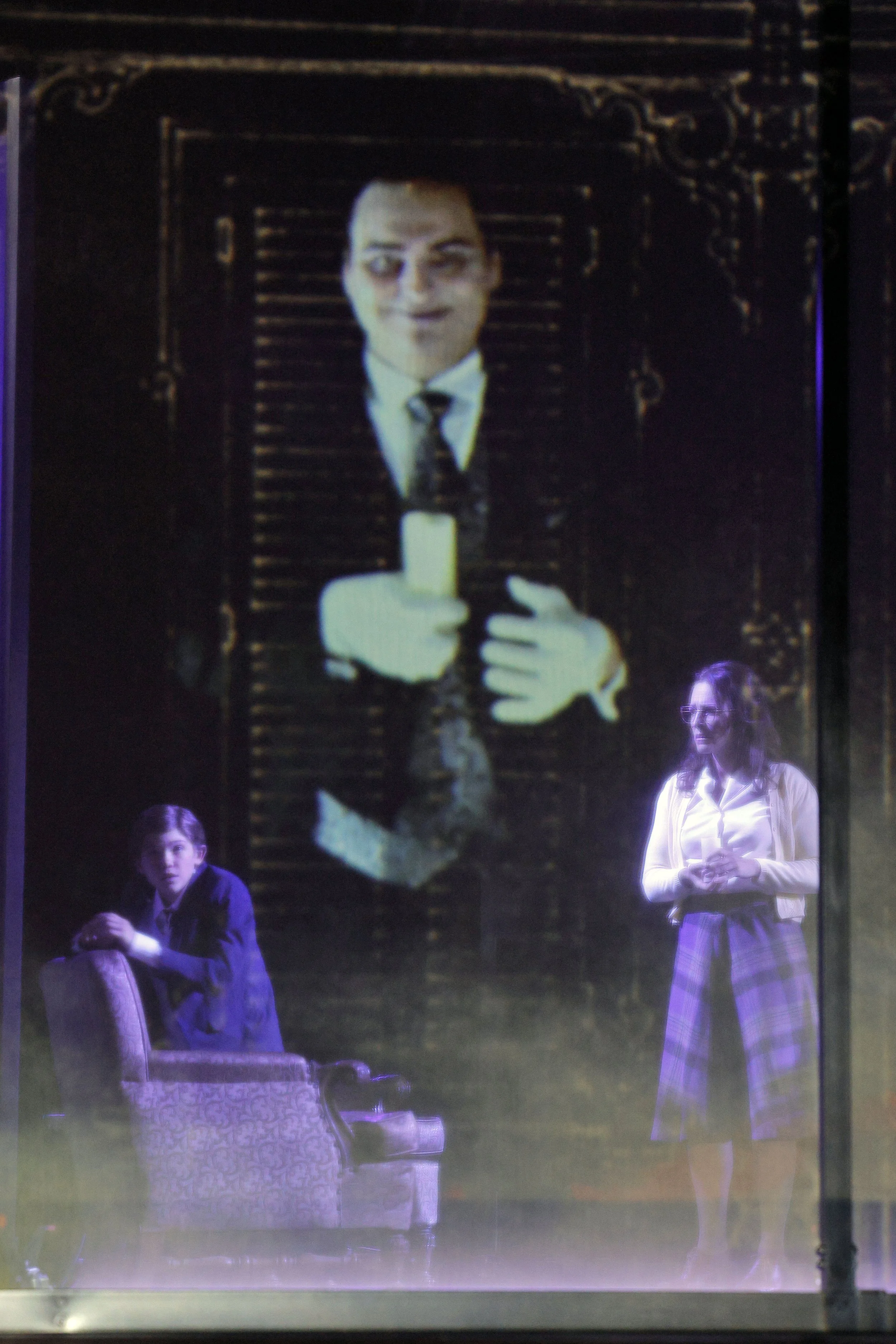 Two actors on stage, a man in a suit and a woman in a plaid skirt, standing in front of a large projection of a man holding a candle.