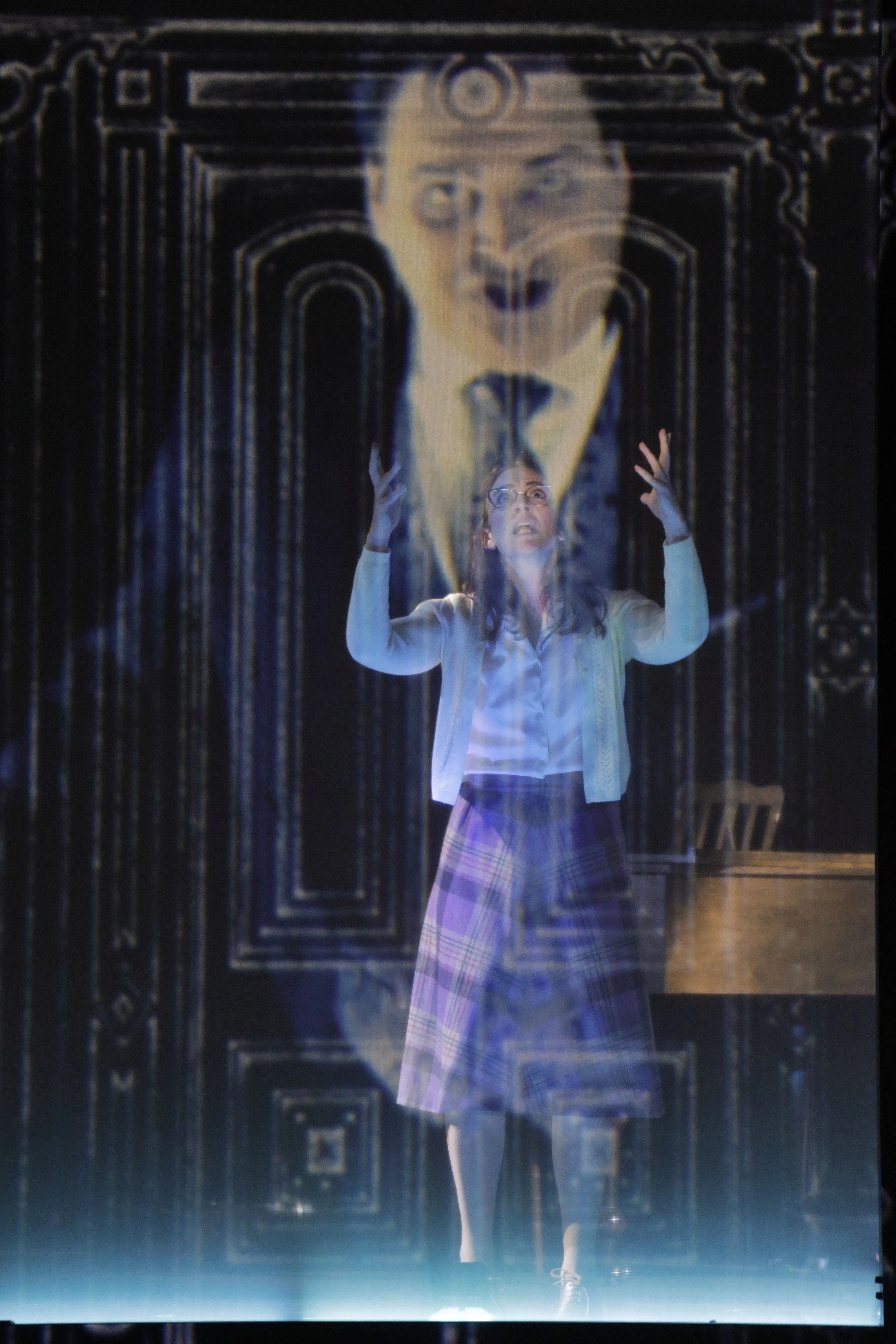 A person with glasses and long hair, wearing a white shirt and plaid skirt, appears to be looking up and gesturing with hands, overlaid with a large, ghostly face of a man with glasses and long hair, set against a dark, ornate background.