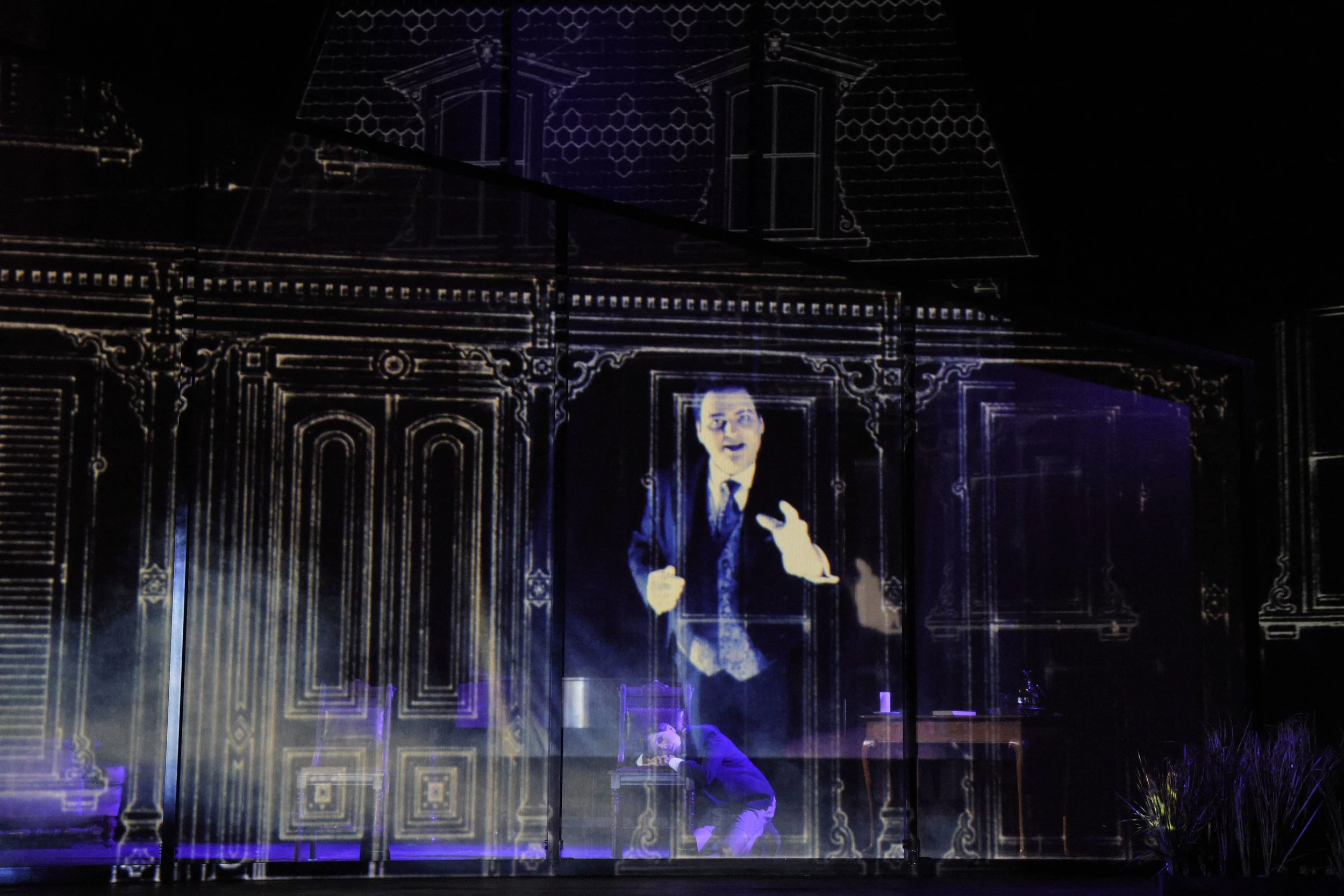 A projection of a man in a suit speaking, displayed on a backdrop of a house with ornate architectural features. The projection includes a smaller figure of a person kneeling in front of the projection.