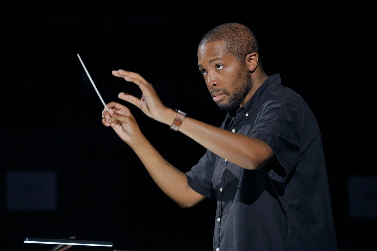 A man conducting an orchestra or choir, holding a conductor's baton, with a focused expression, wearing a dark shirt against a black background.