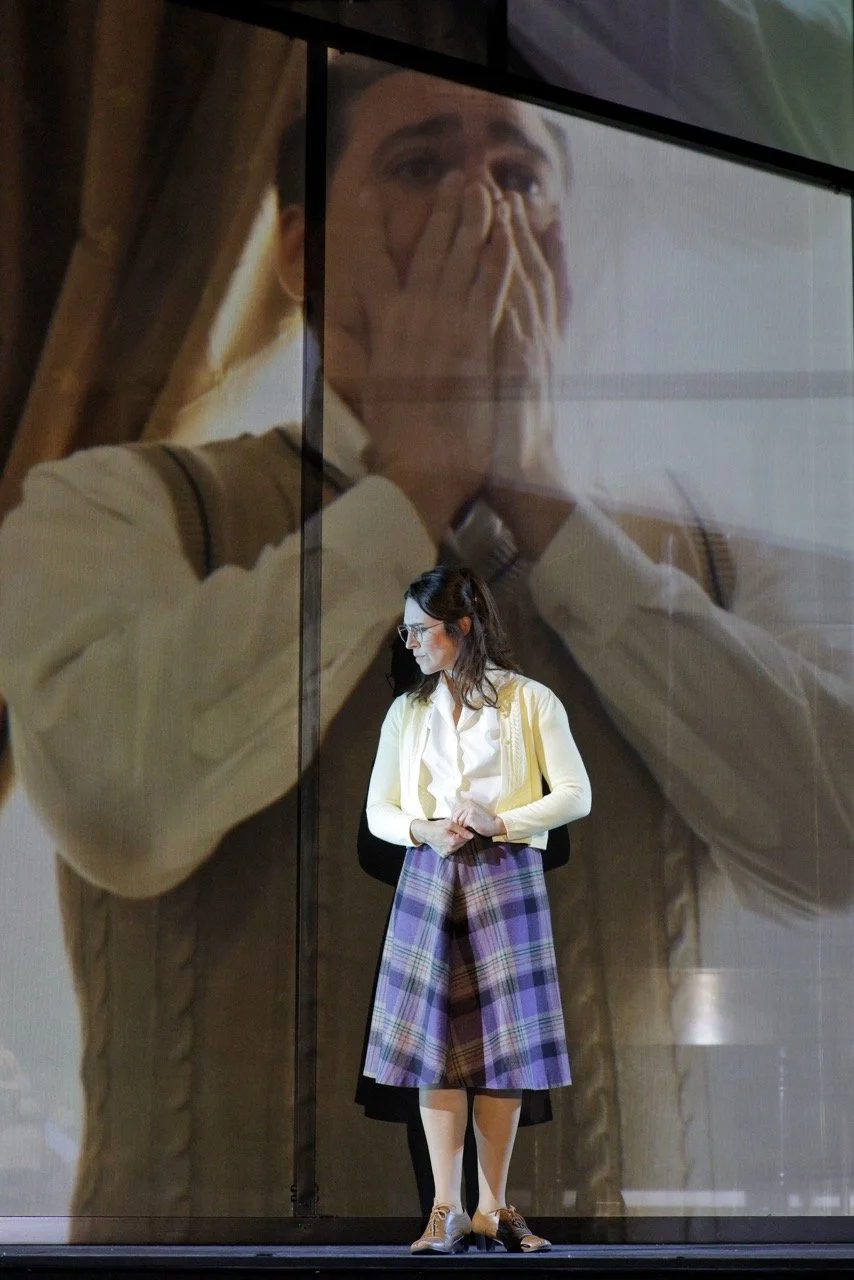 A woman standing indoors in front of a large glass display shows her reflection looking surprised or shocked, with her hands covering her face.