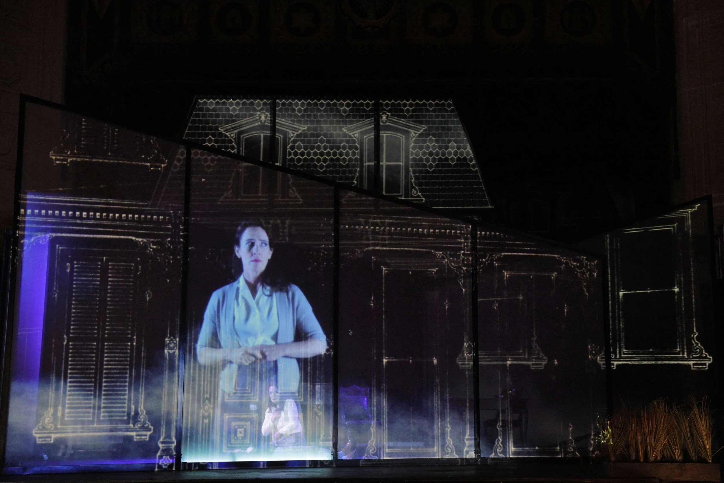 A projection of a woman in a white shirt with folded arms standing in front of a historical building with ornate wooden doors and decorative details, displayed on glass panels.