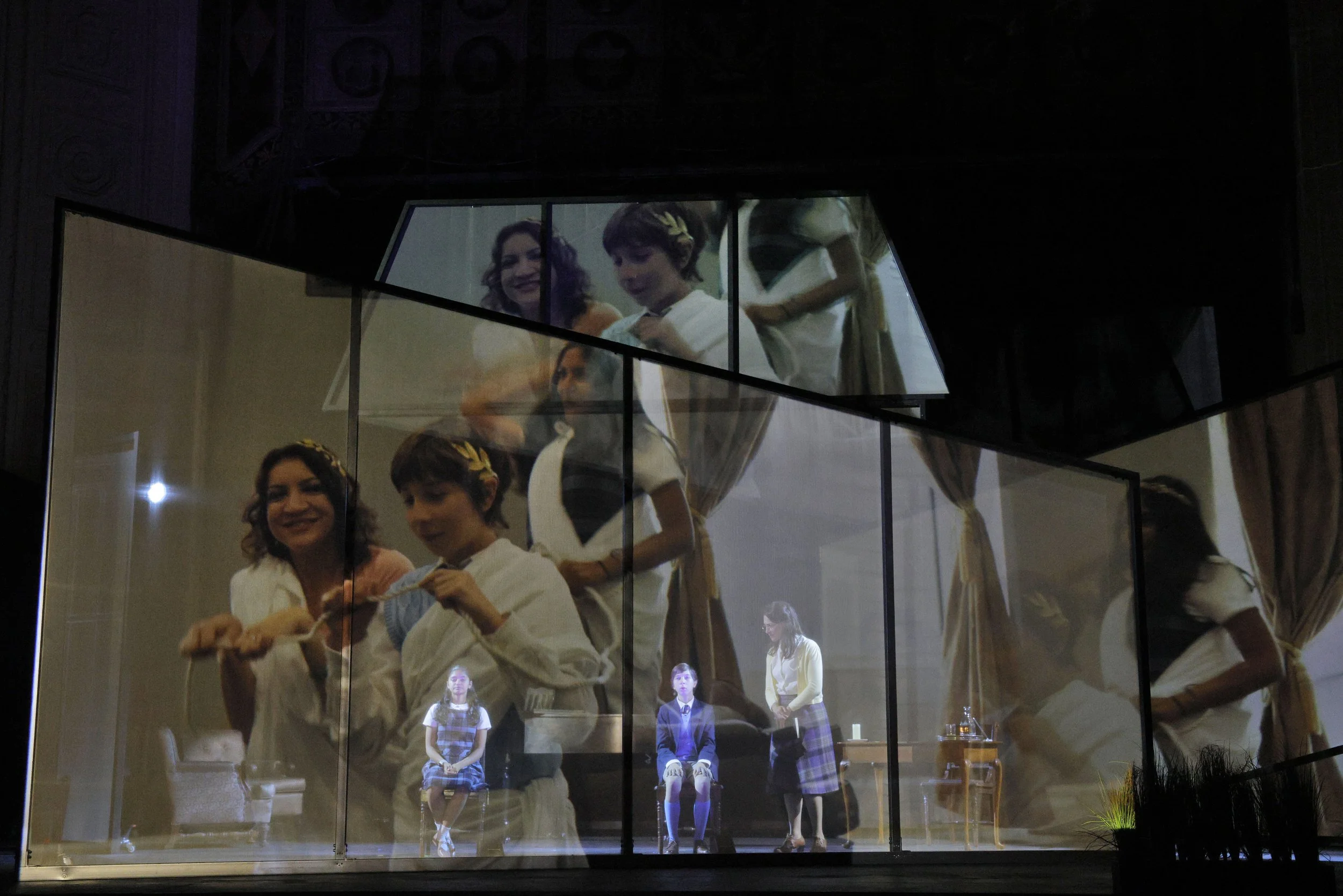 People on stage behind a glass window, performing or rehearsing, with some holding papers, and others seated or standing, with a dark background and stage lights.