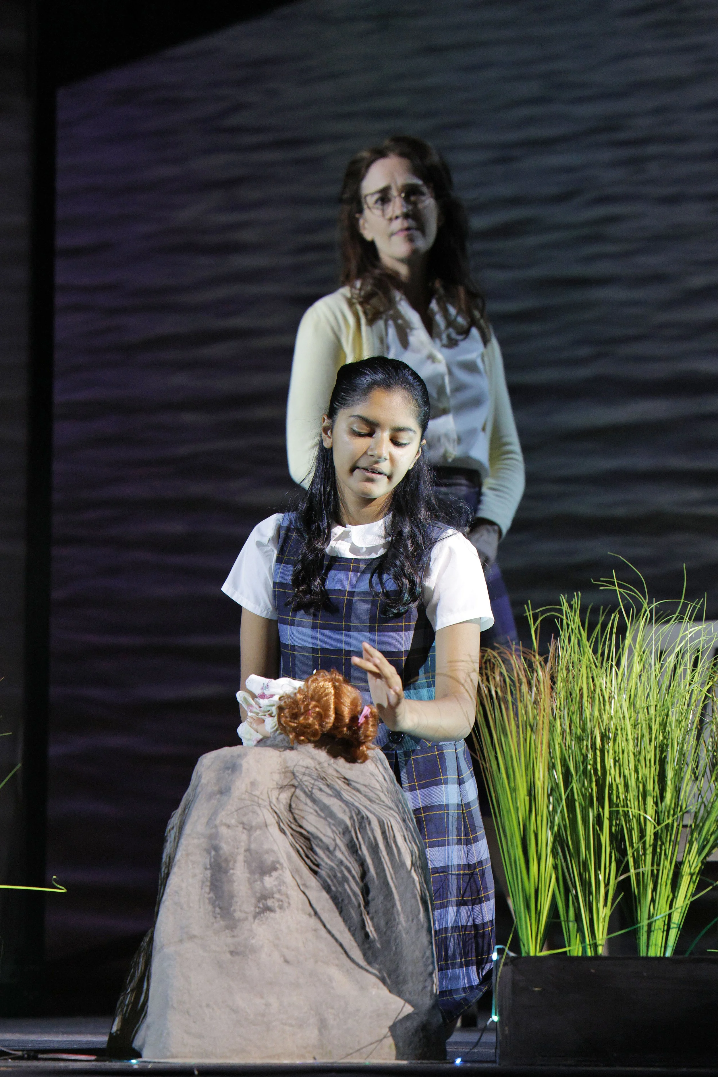 Two young girls, one with long dark hair and the other with glasses and brown hair, are performing on stage with a backdrop that resembles water. The girl in front is wearing a school uniform and holding a puppet or doll with curly red hair, while th