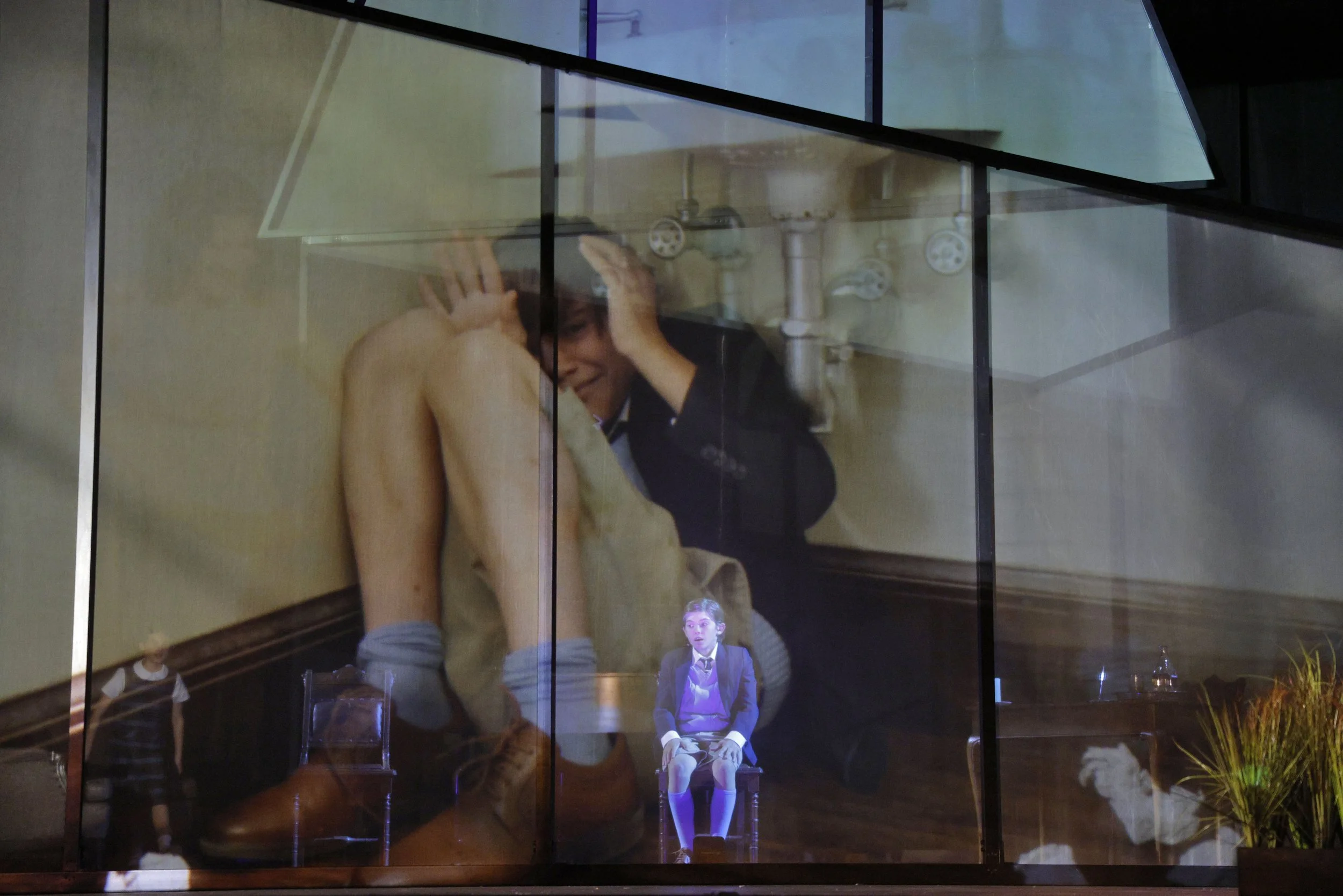A reflection of a woman with short hair, wearing a bra, holding her head with her hands inside a glass booth. In the background, sitting on a chair, is a person in a purple blazer and tie, with a surprised expression.