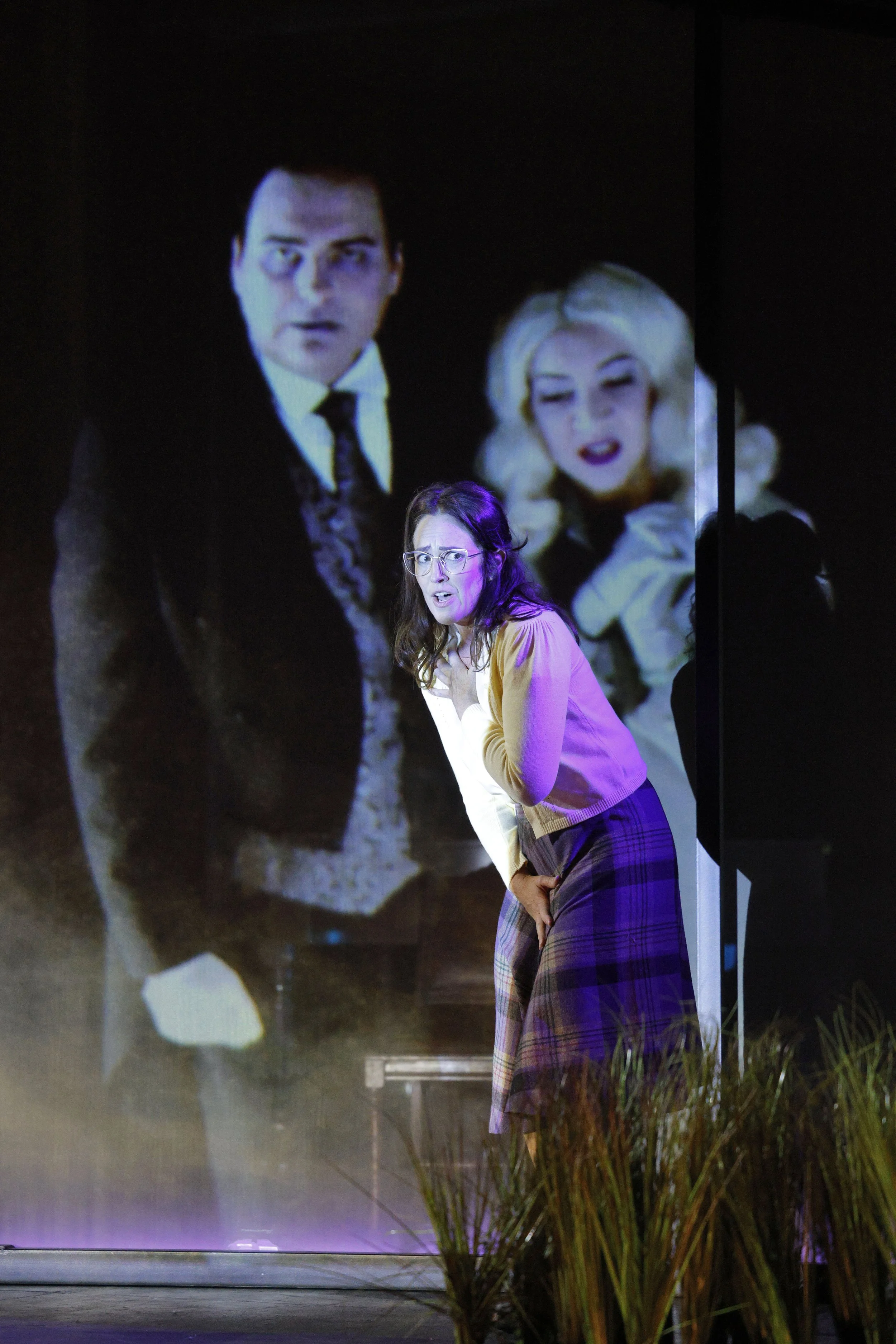 A woman with glasses and long brown hair, wearing a yellow top and plaid skirt, appears surprised or concerned, standing indoors in front of a reflective surface that shows a large projection of a man in a suit with a serious expression and a woman w