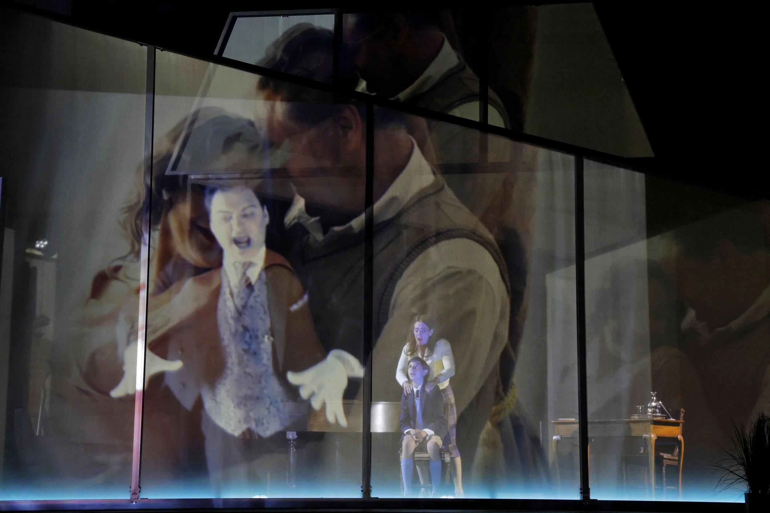 Multiple projections of people on a glass wall, including a woman, a man, and two children, with reflections and lighting creating a layered visual effect.