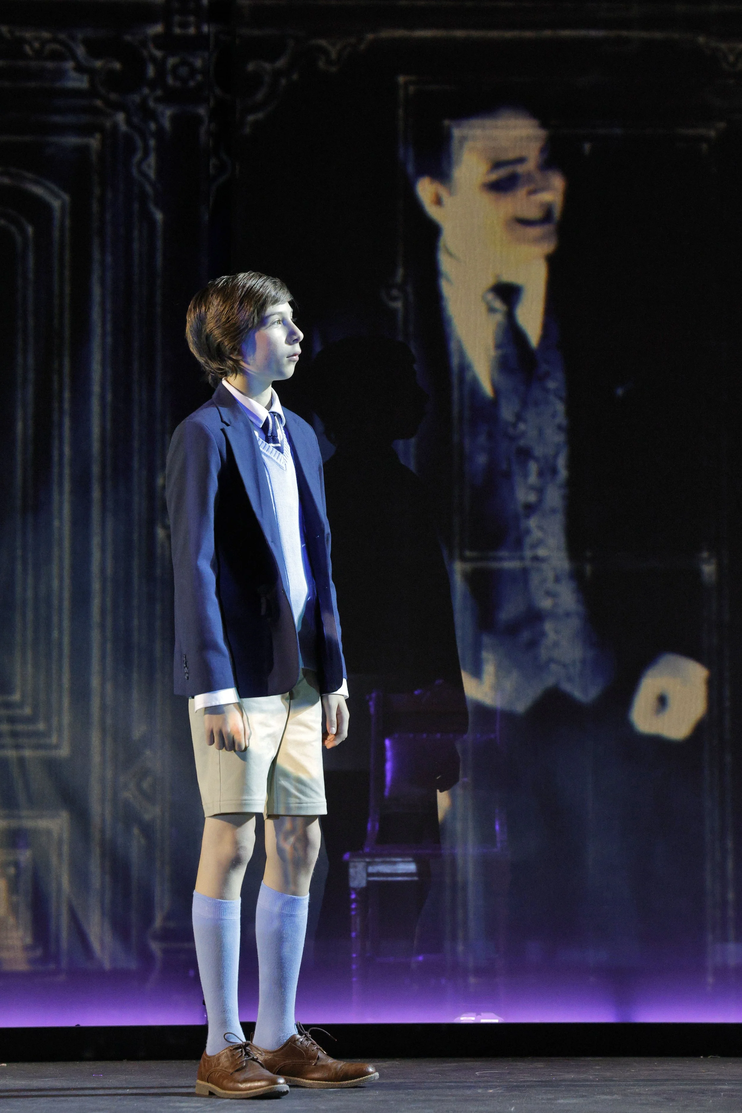 A young man with medium-length brown hair, dressed in a blazer, sweater vest, collared shirt, and beige shorts, standing on a stage with a large screen displaying his image in the background.