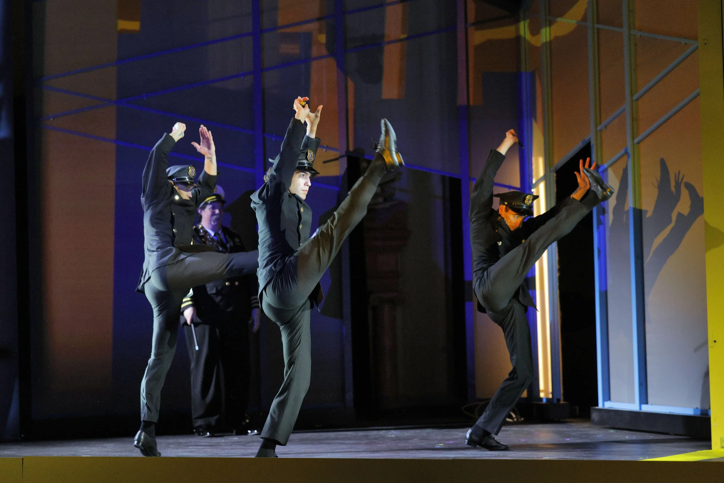 Three performers dressed as police officers are doing high kicks on a stage, with a colorful backdrop and stage lighting.