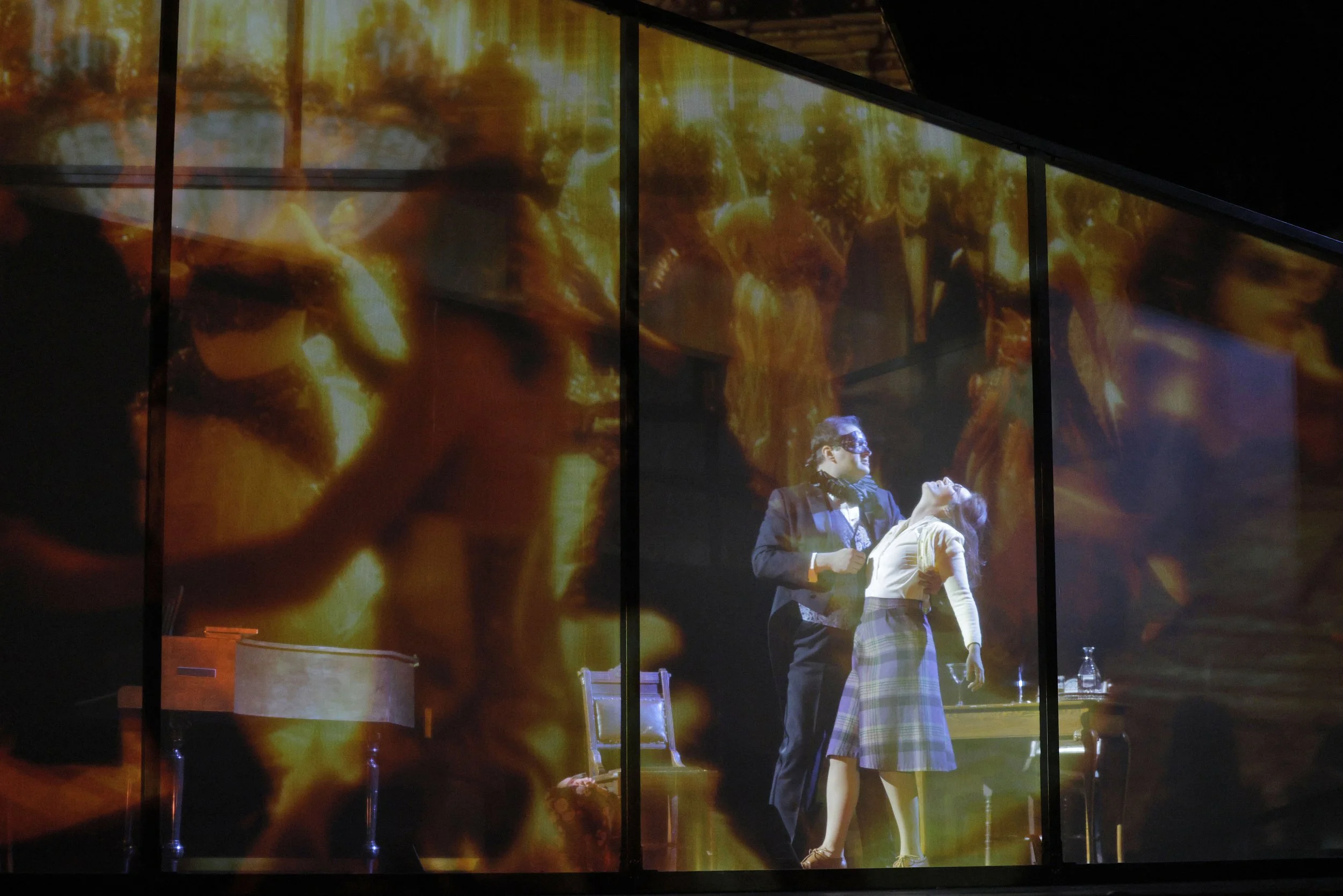 A theatrical scene with two people, one man and one woman, standing close together on stage with a background of fiery flames and flames projected on glass. The man is wearing a tuxedo and the woman is in a plaid skirt and white sweater.